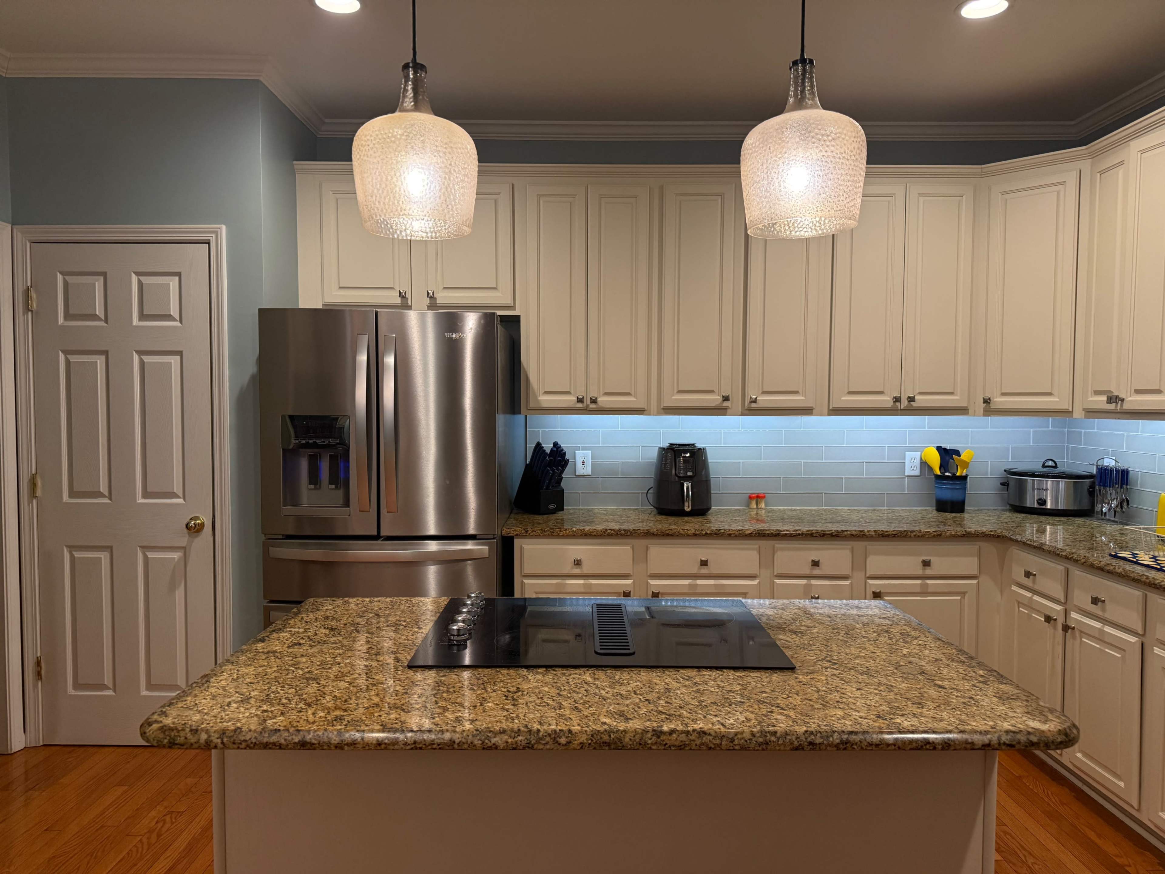 A modern kitchen features light-colored cabinetry, a large granite island with a cooktop, stainless steel appliances, and pendant lighting.
