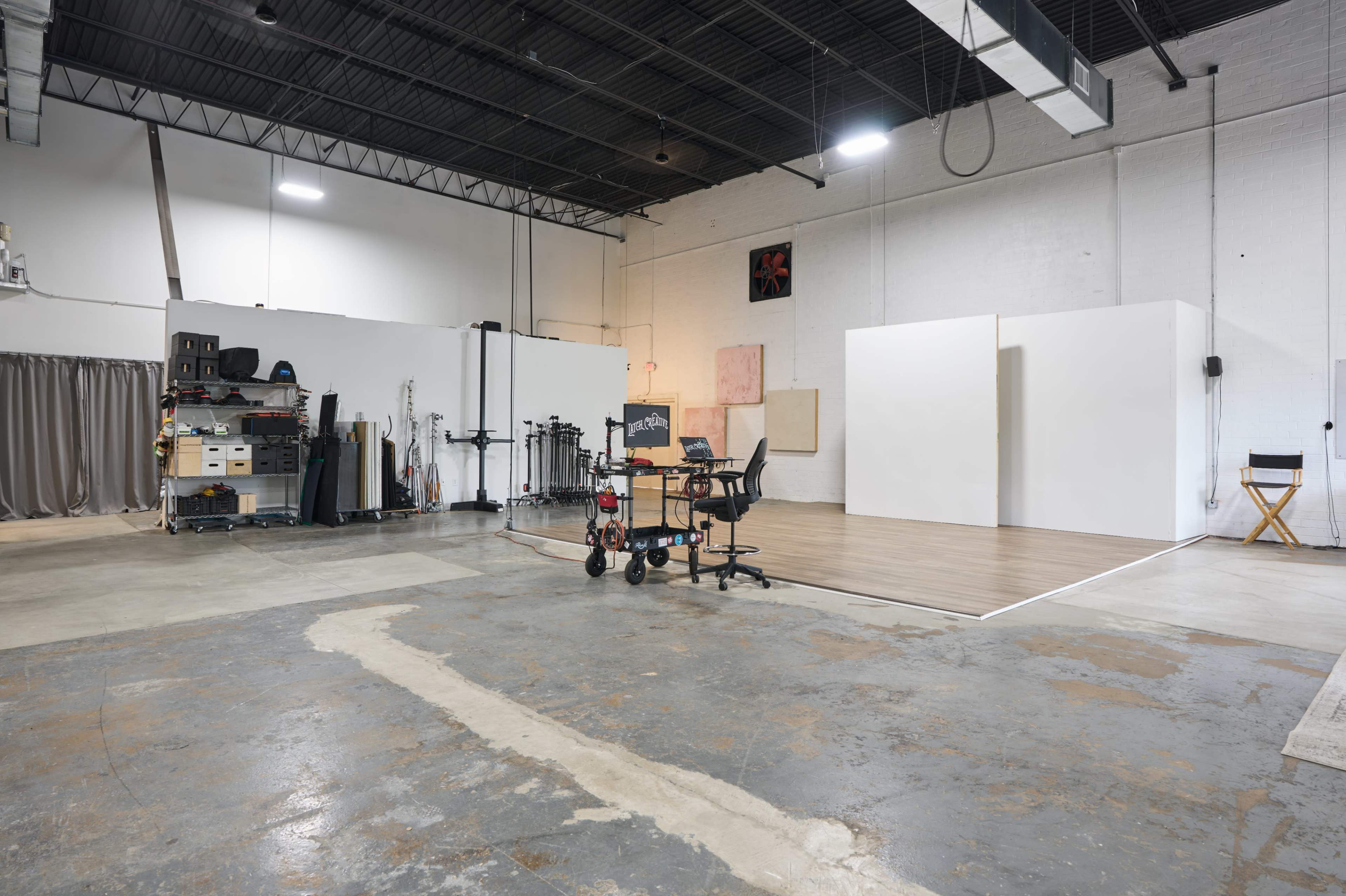 The image shows a spacious studio with white walls, a wooden floor area, and various equipment including chairs and a shelving unit along one side.