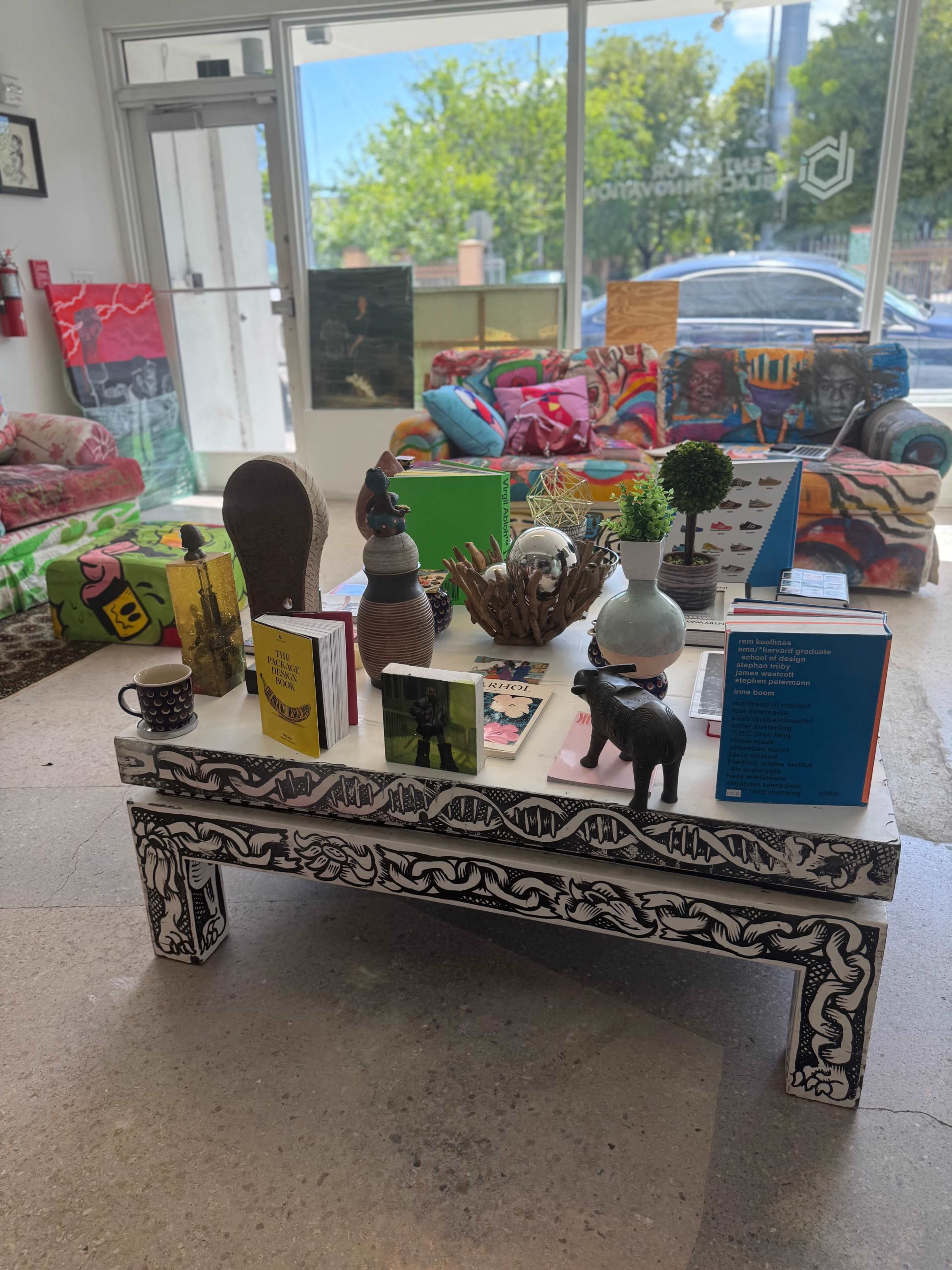 The image shows a vibrant art space featuring a decorated table with books and decorative objects, surrounded by colorful sofas.