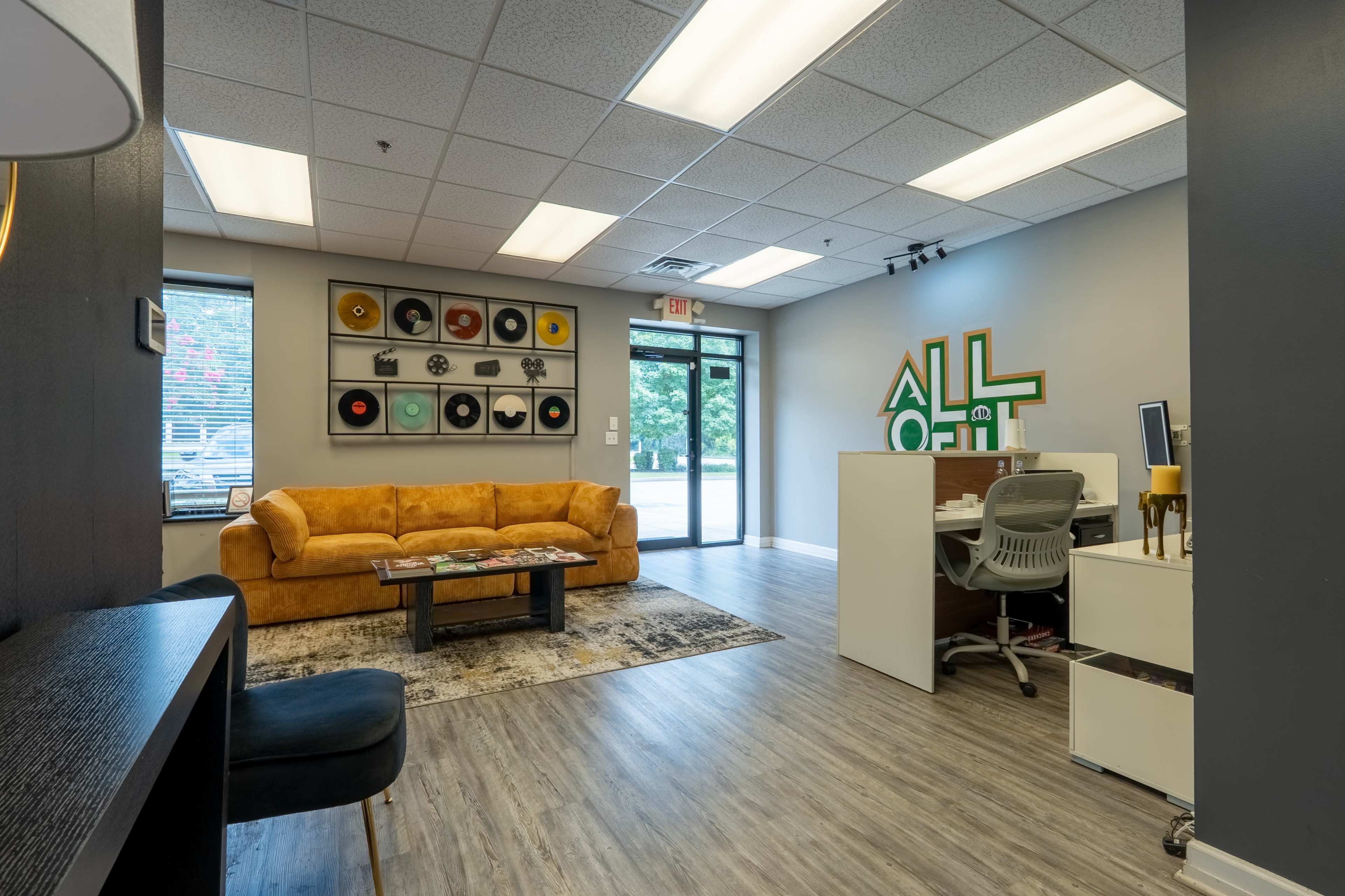 The image shows a modern office space featuring a yellow couch, a coffee table, a desk with a chair, and a wall adorned with framed records and a vibrant "ALL OUT" sign.