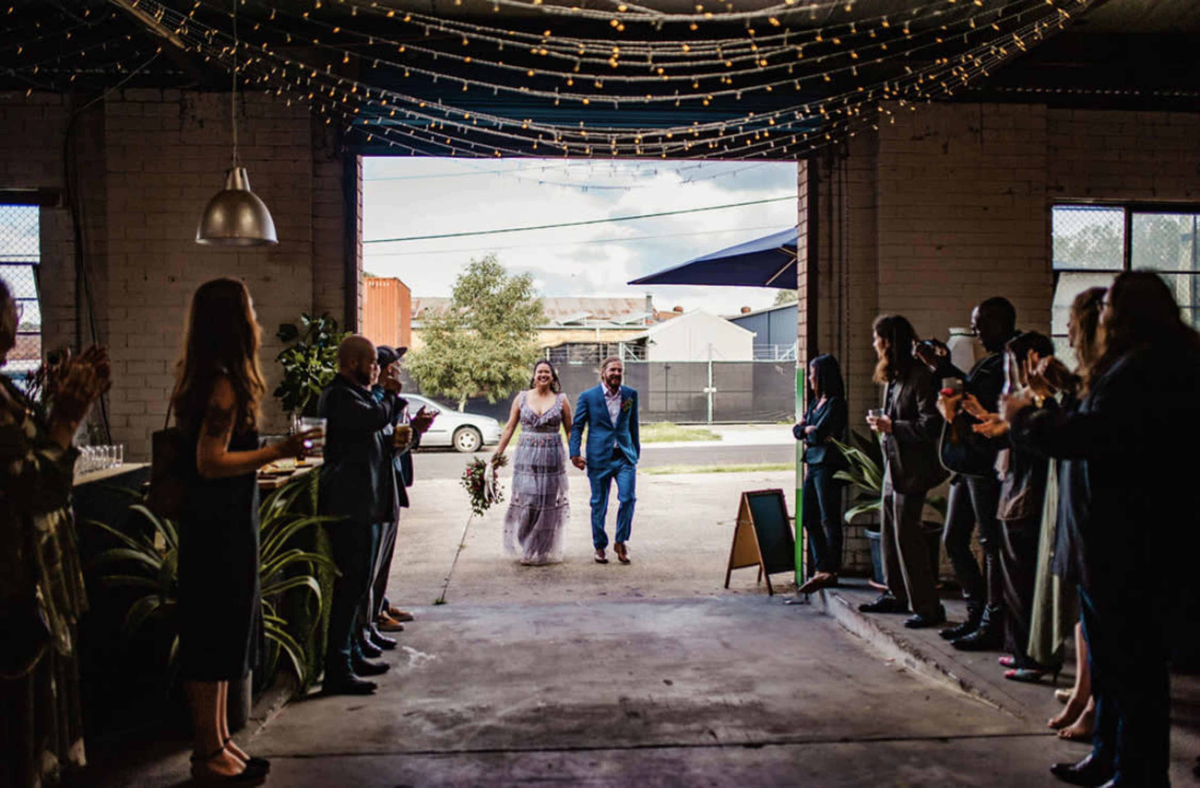 A bride and groom walk through an open doorway, greeted by guests who are applauding in a decorated industrial space.