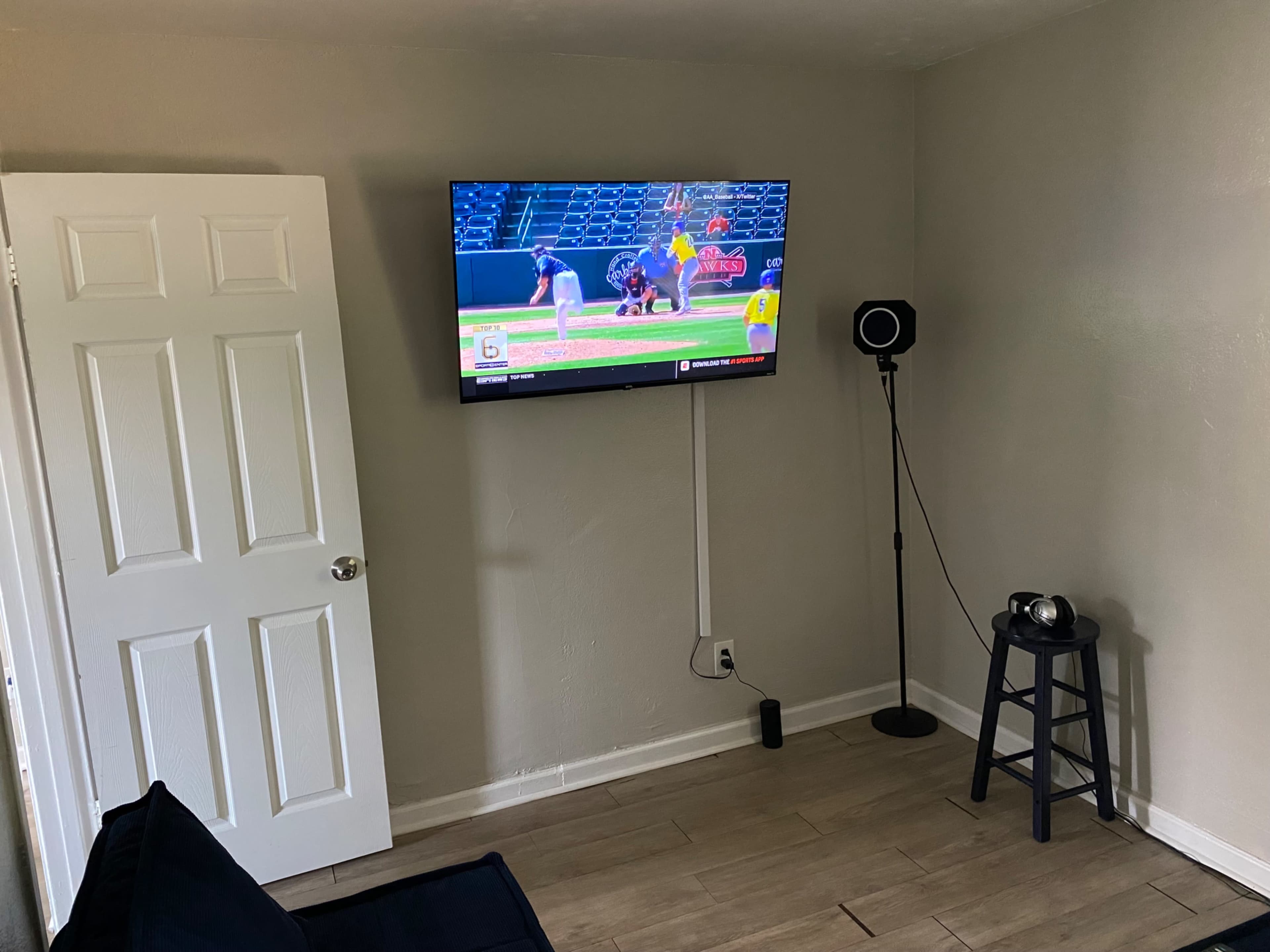 A television displaying a baseball game is mounted on a wall opposite a small seating area with a chair and a floor lamp.