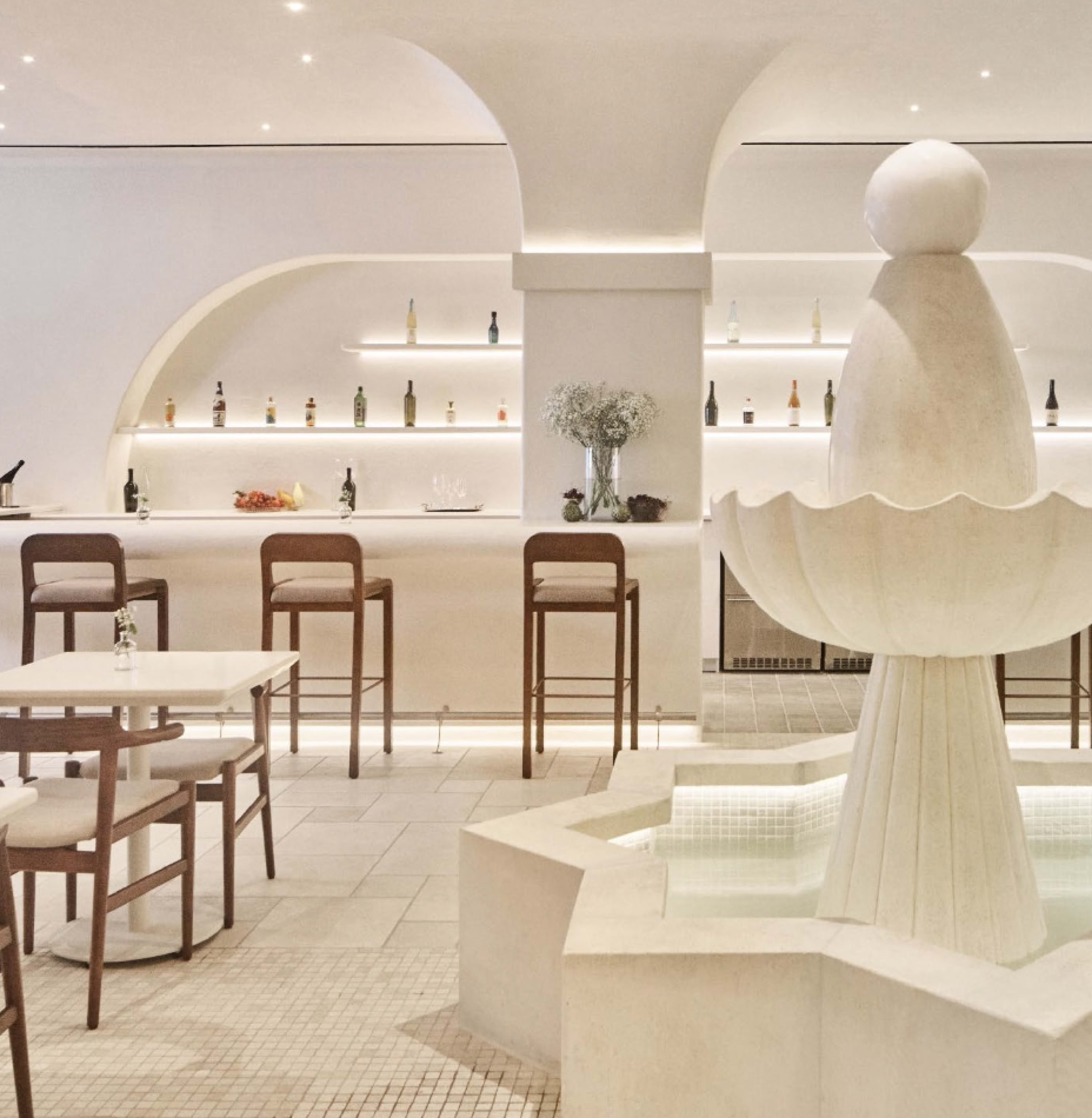 The image features a modern, minimalist restaurant interior with a central fountain surrounded by bar stools and tables.