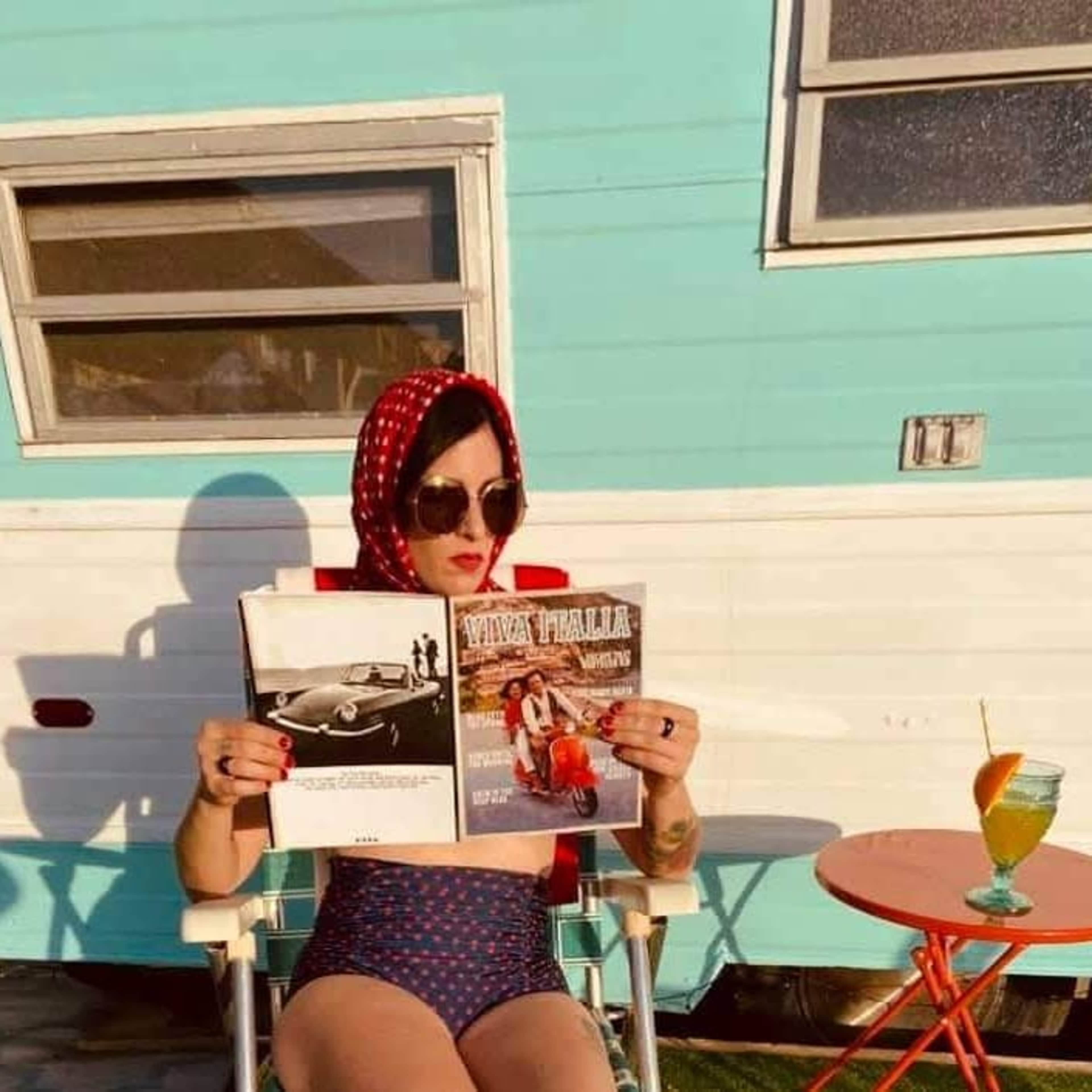 A woman sits in a chair outside a vintage trailer, reading a magazine while holding a colorful drink.