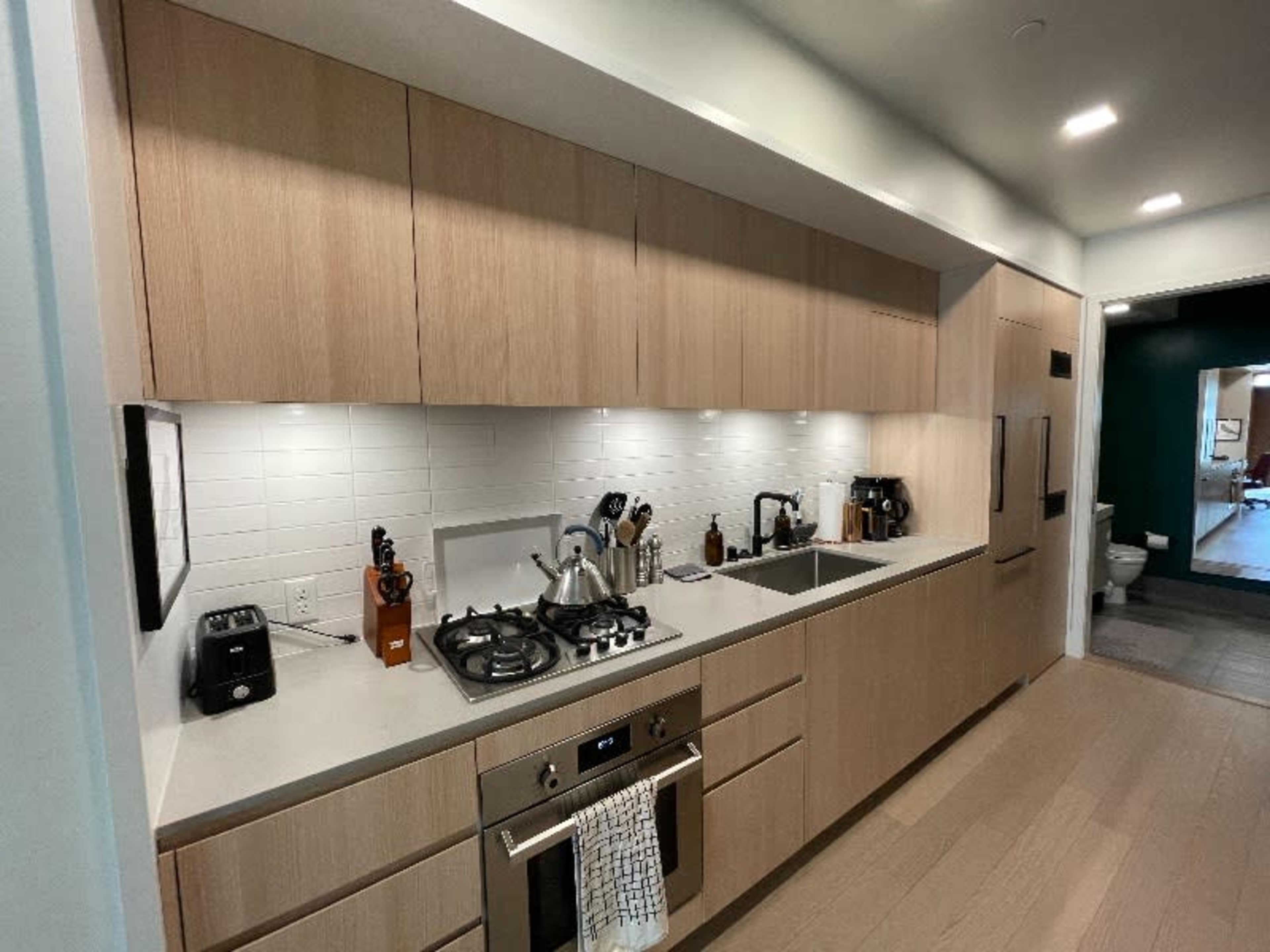 A modern kitchen features light wood cabinetry, a stainless steel stove, a double basin sink, and various kitchen appliances neatly arranged along the countertop.