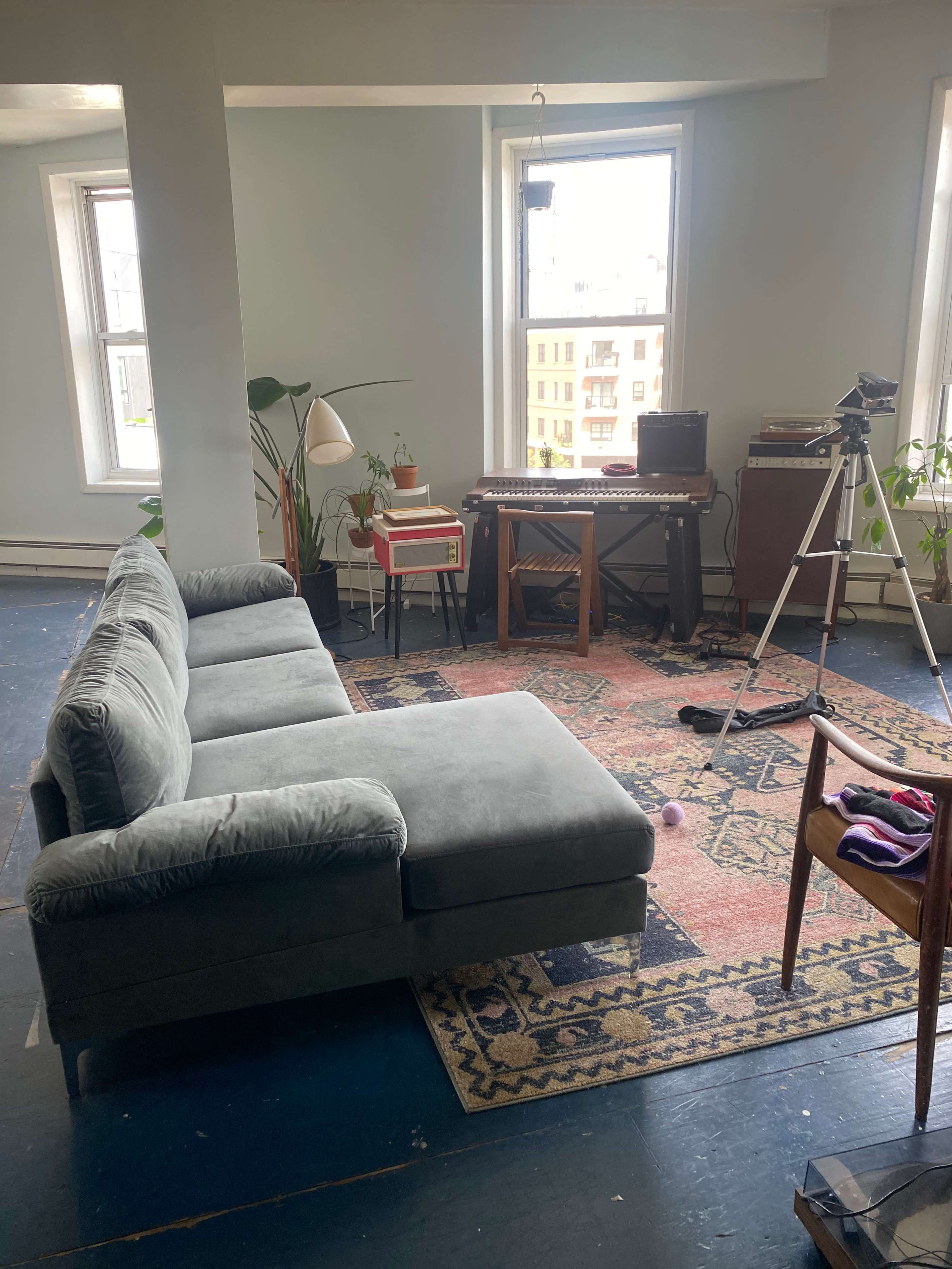 A gray sectional sofa is positioned on a rug in a light-filled room, with a plant, a small red side table, a keyboard on a wooden stand, and a tripod set up nearby.