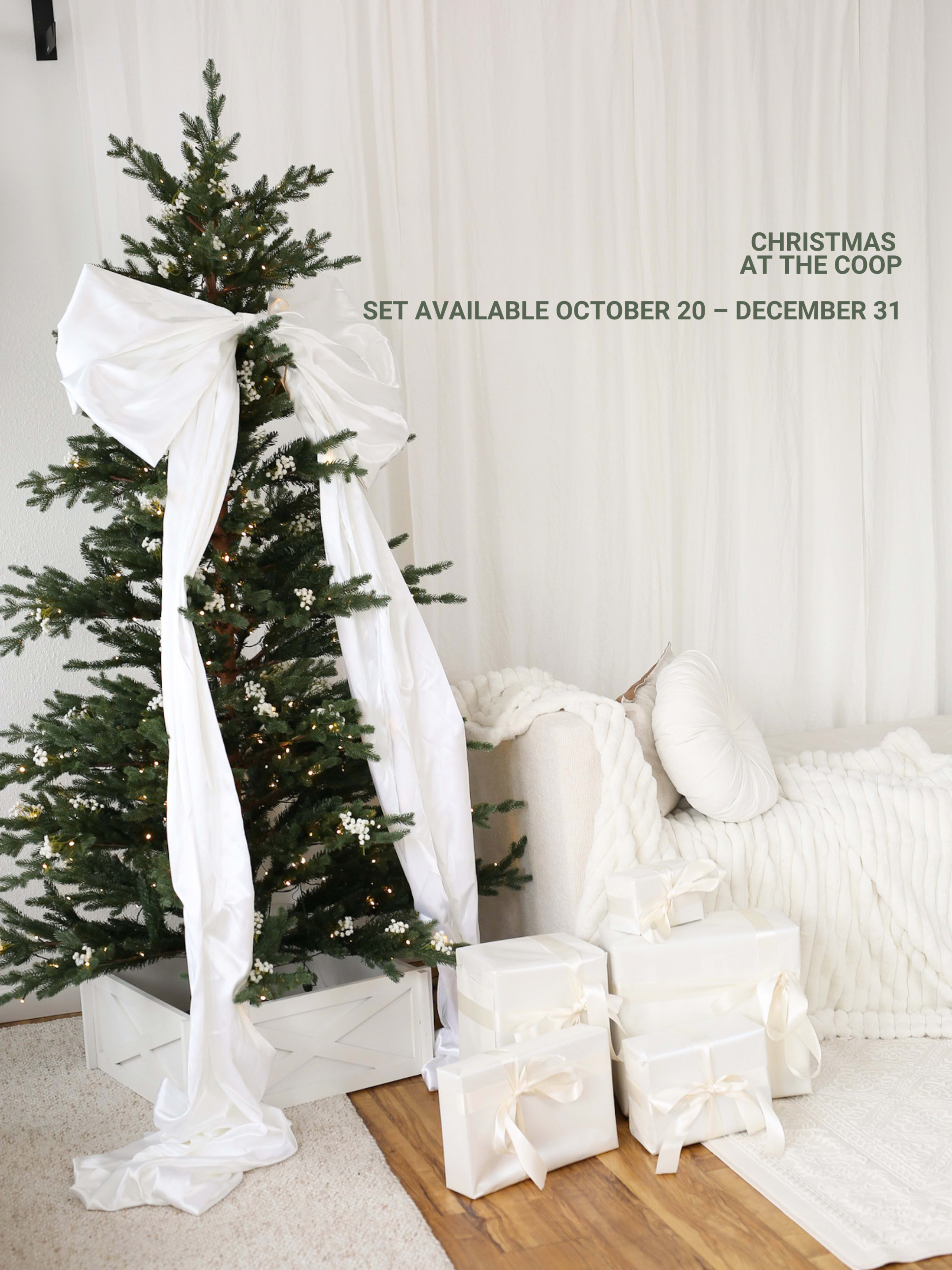 A Christmas tree adorned with a large white bow stands next to a cozy sofa, surrounded by elegantly wrapped packages.
