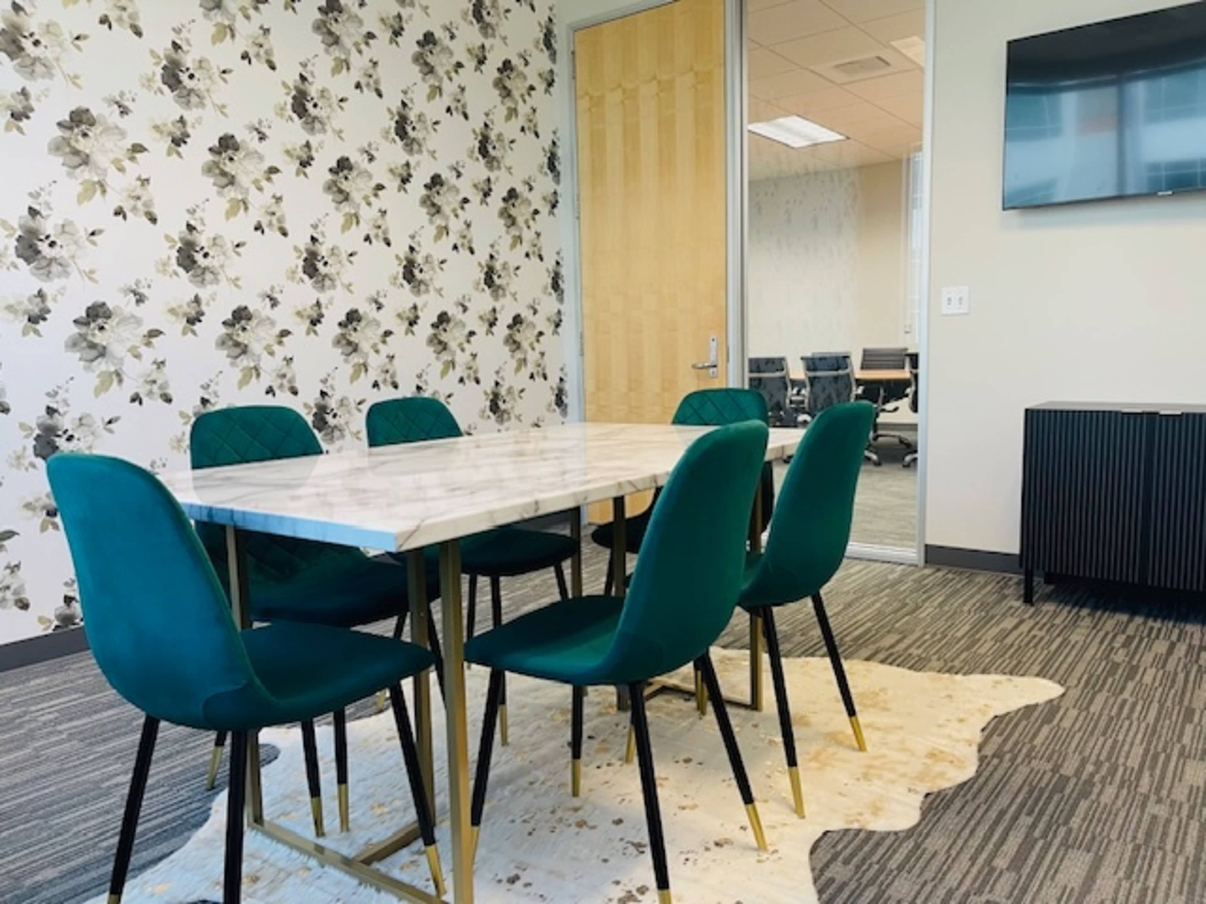 A conference room features a marble-top table surrounded by six green chairs on a patterned carpet, with a TV mounted on the wall and a glass-walled meeting area visible in the background.