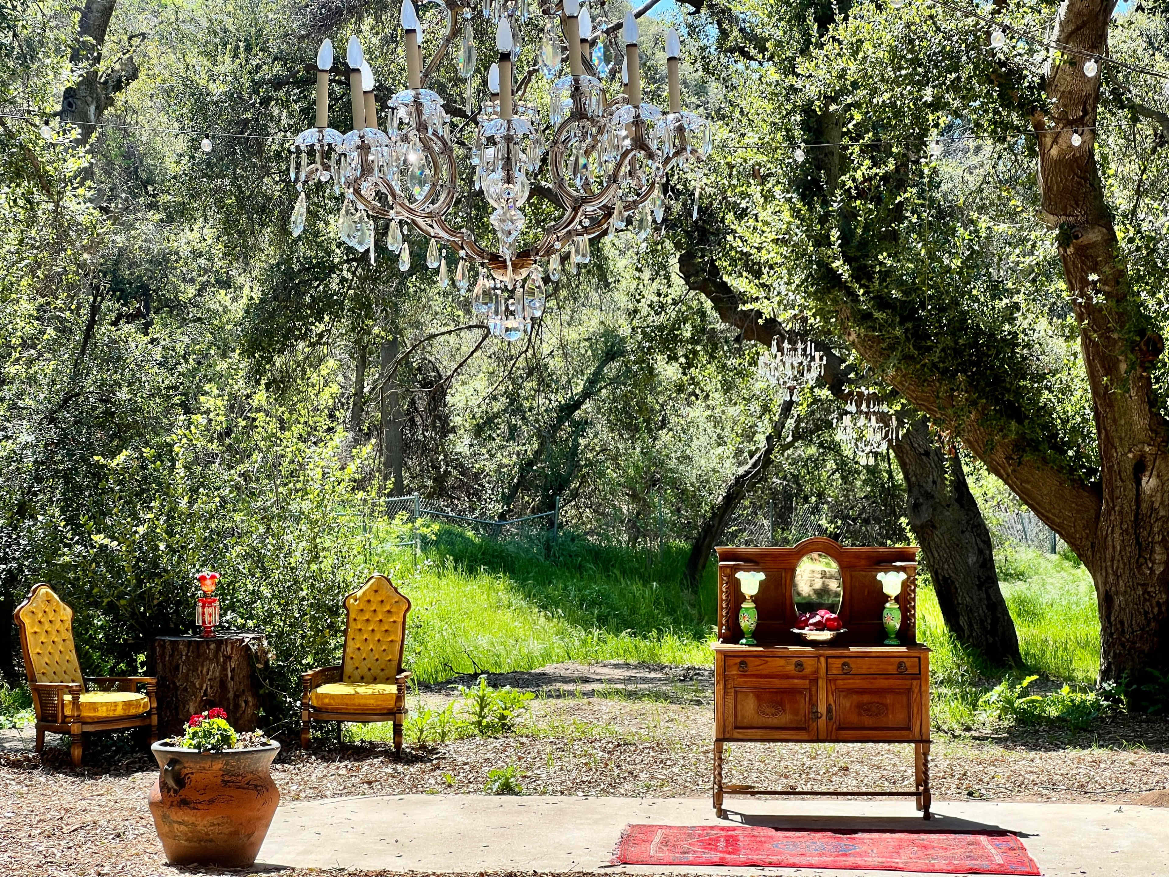 The scene features a rustic outdoor setting with a wooden table and two yellow chairs under a chandelier hanging from the trees, surrounded by greenery.