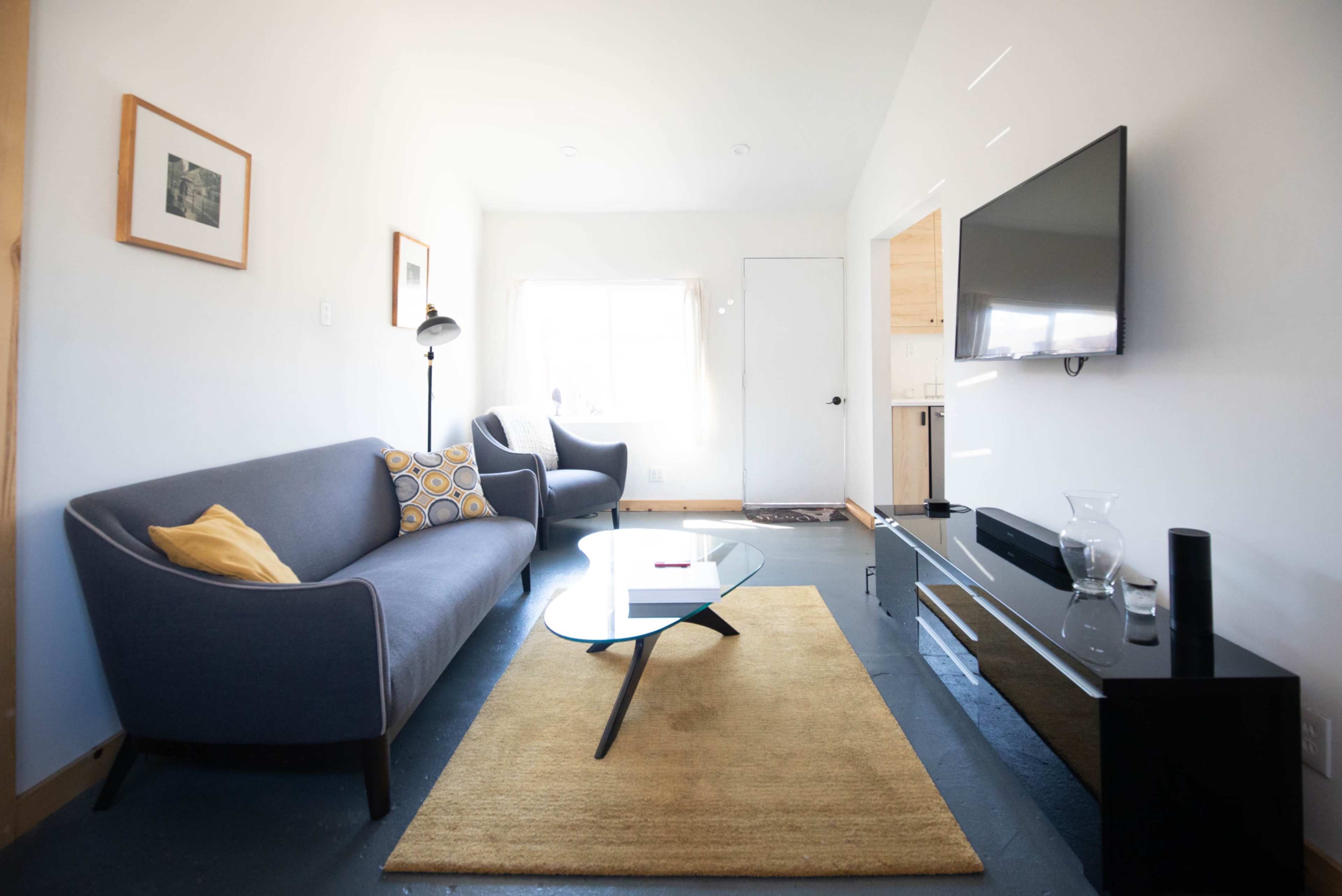 The image shows a living room with a gray sofa, a round coffee table, a flat-screen TV mounted on the wall, and a beige area rug.