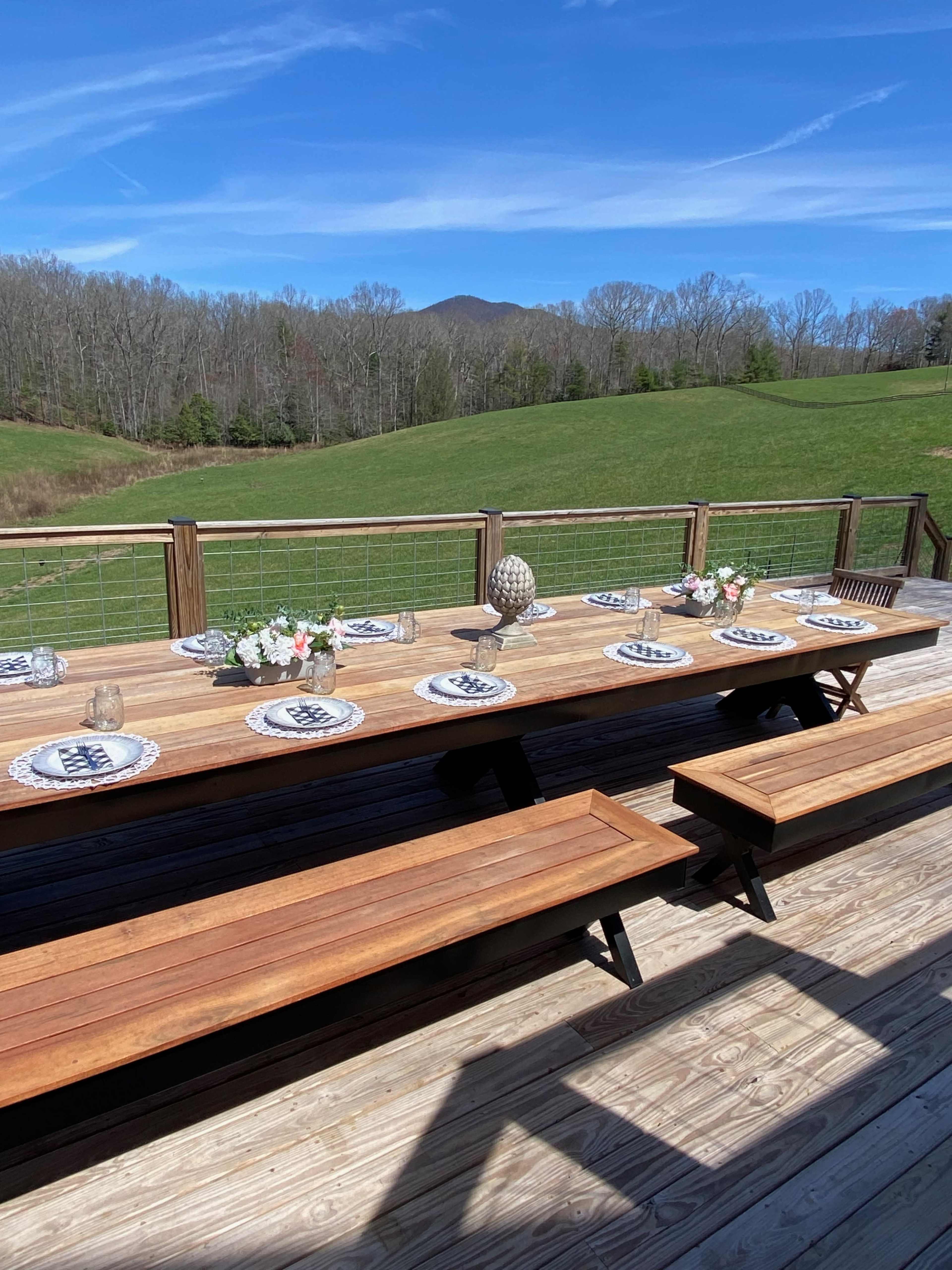 A long wooden table is set with decorative plates and floral arrangements on a deck overlooking a green field and distant mountains.