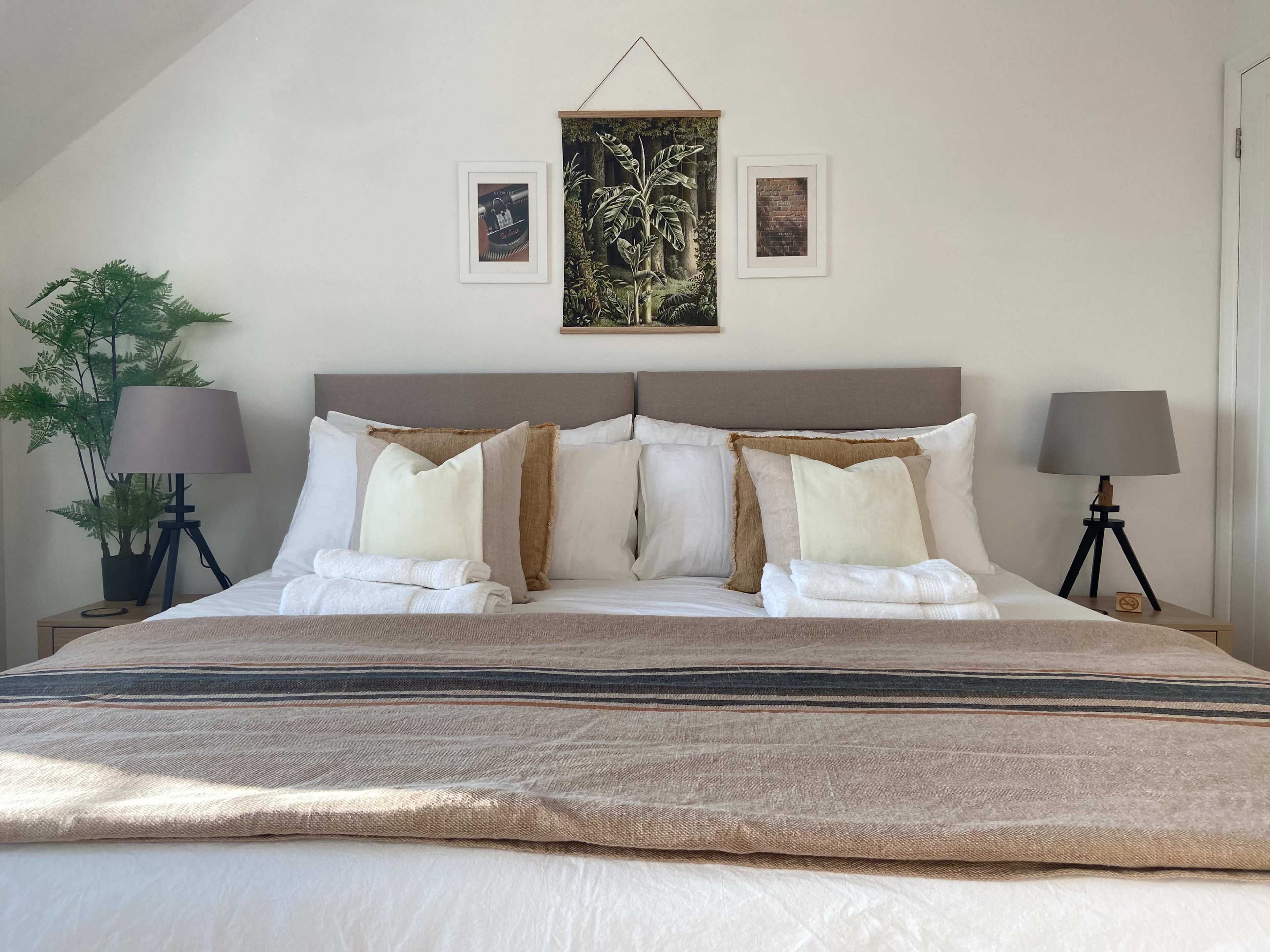 A neatly arranged bedroom with a large bed adorned with various pillows, a tan throw blanket, and framed artwork hanging on the wall above.