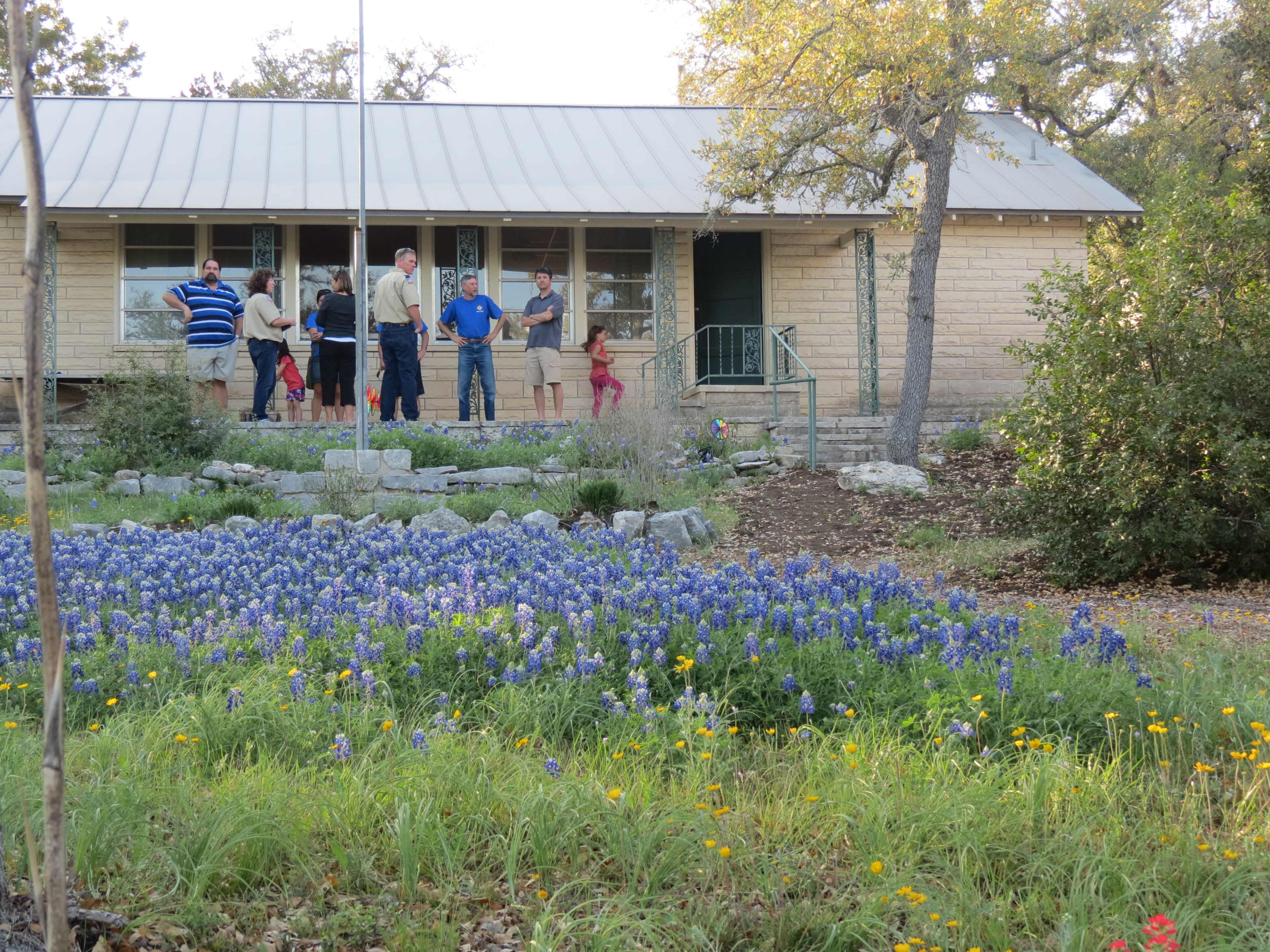 A group of people stands in front of a house surrounded by fields of bluebonnets and other wildflowers.