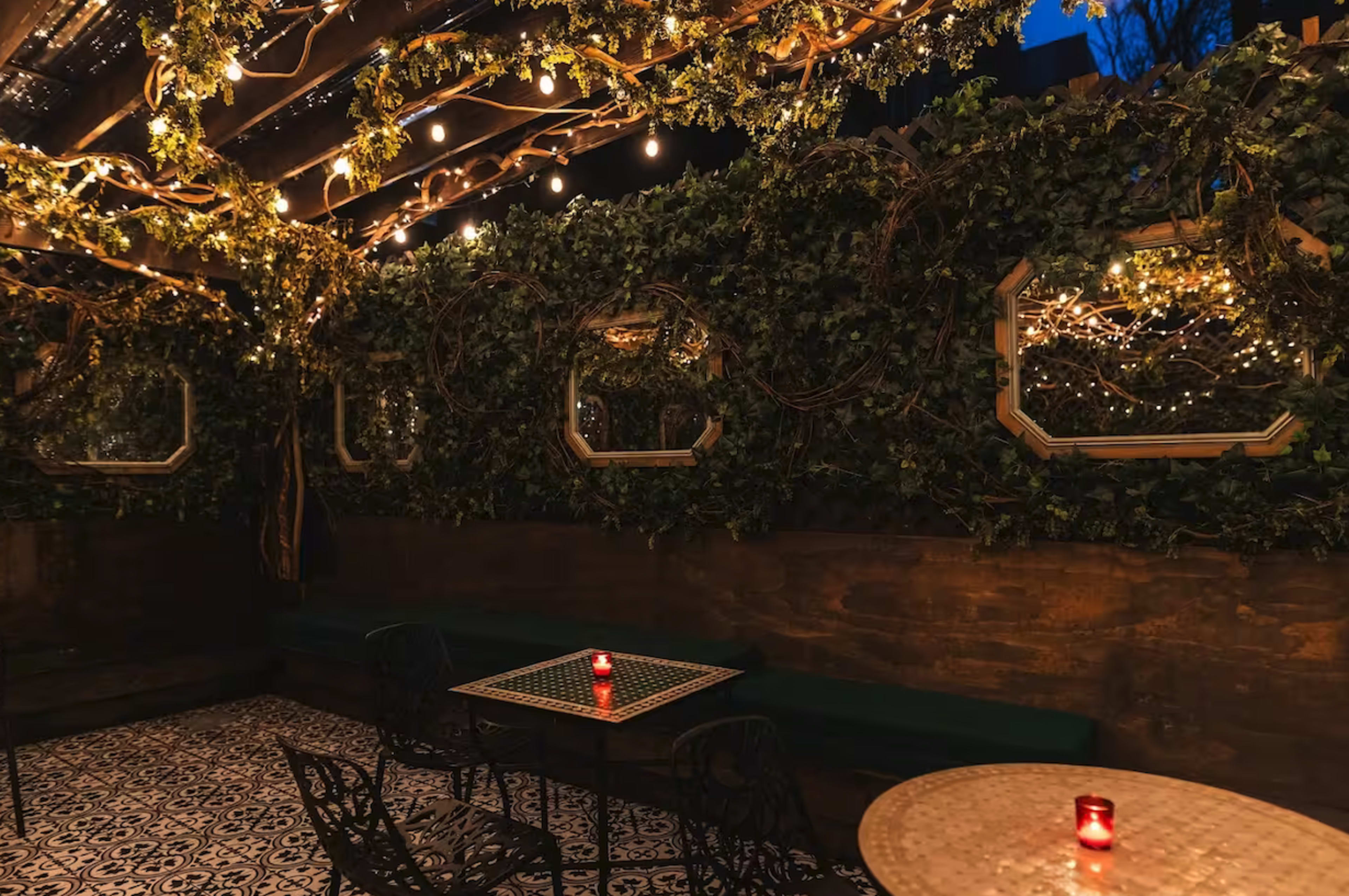 The image shows a cozy outdoor dining area with tables, surrounded by greenery and string lights, creating a warm atmosphere.