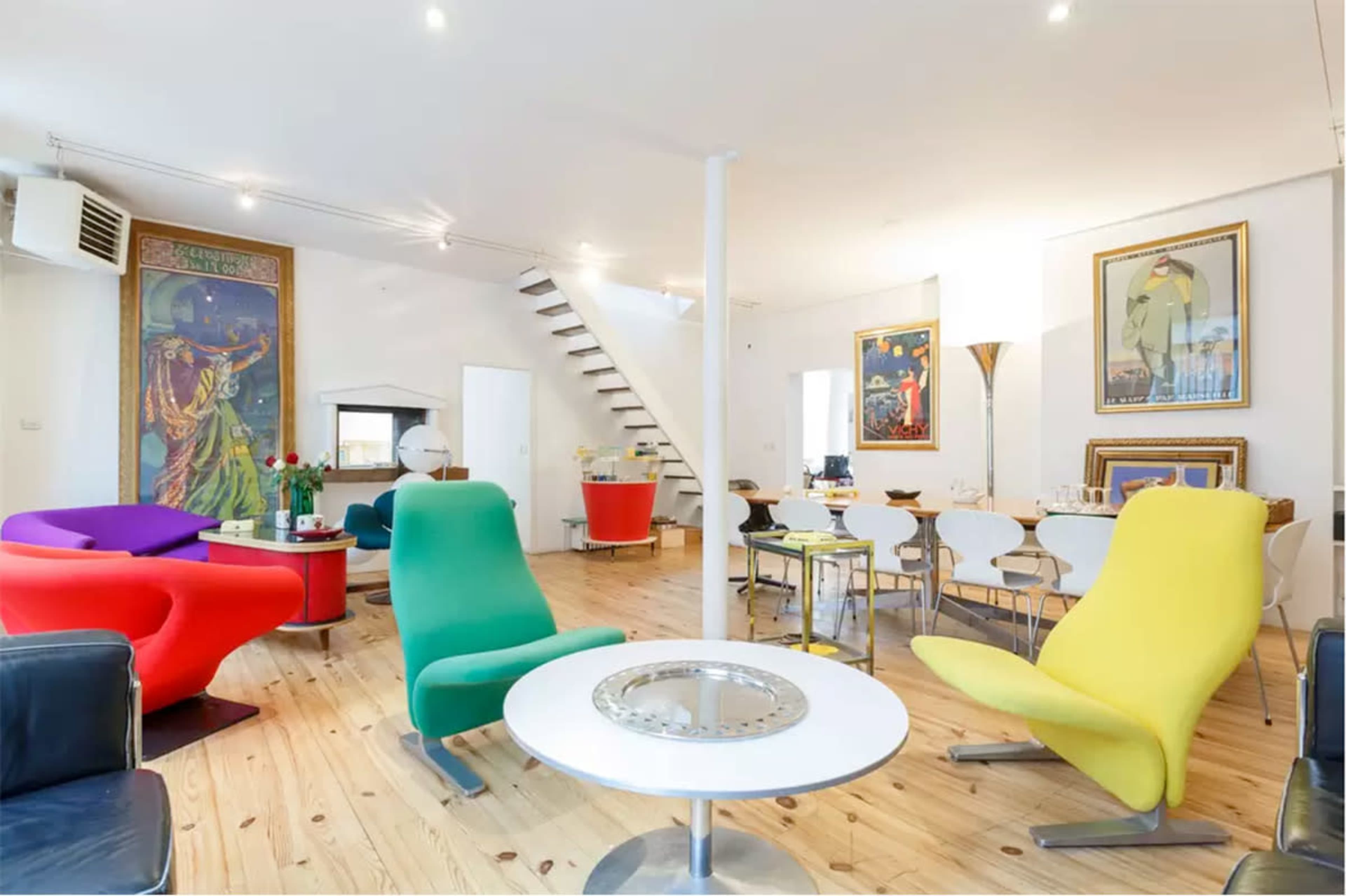 A modern interior space featuring colorful chairs, a round table, and a dining area with white chairs, along with a staircase and framed artwork on the walls.
