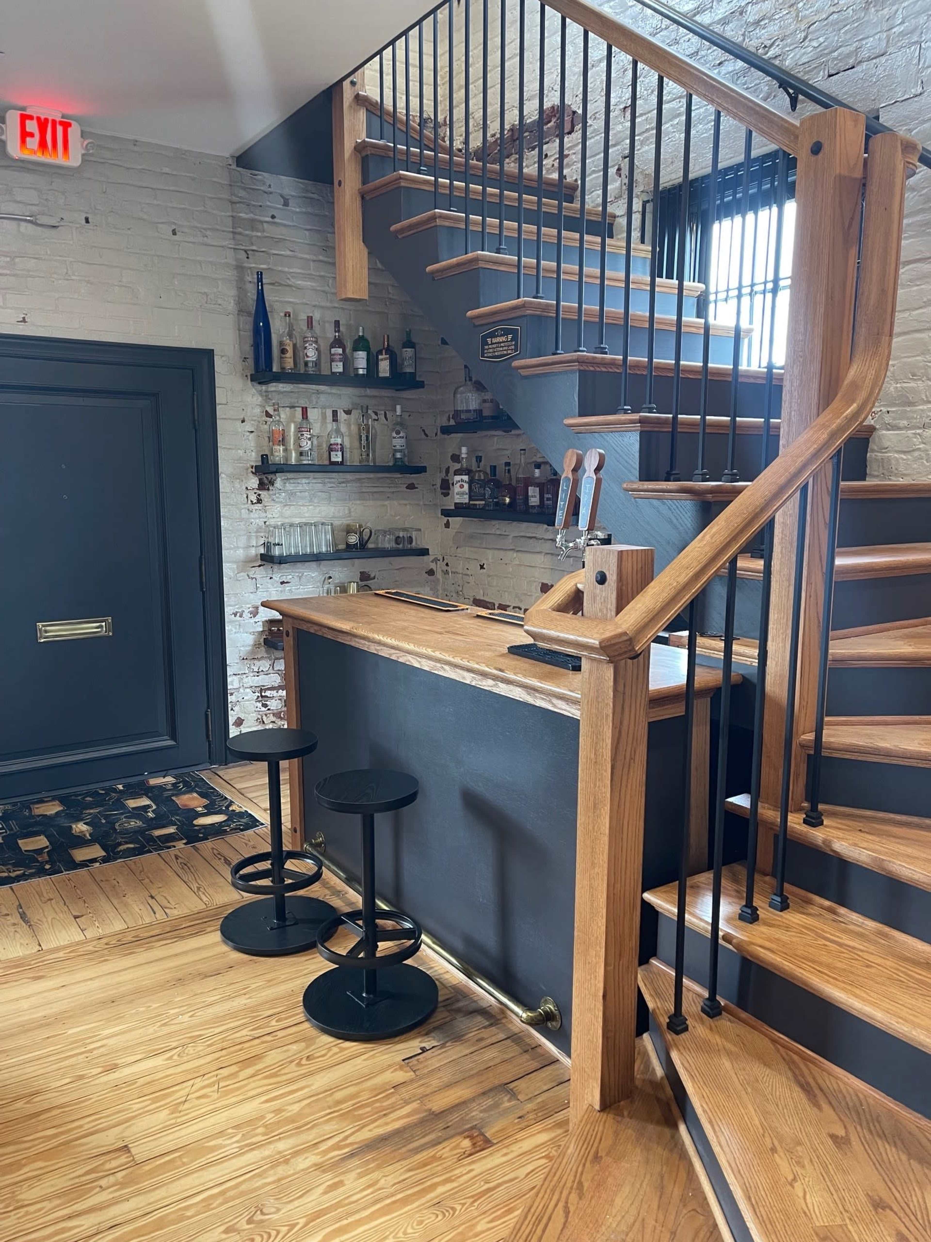 The image shows a wooden staircase leading to an upper level, adjacent to a small bar area with three black stools and shelves displaying various bottles.