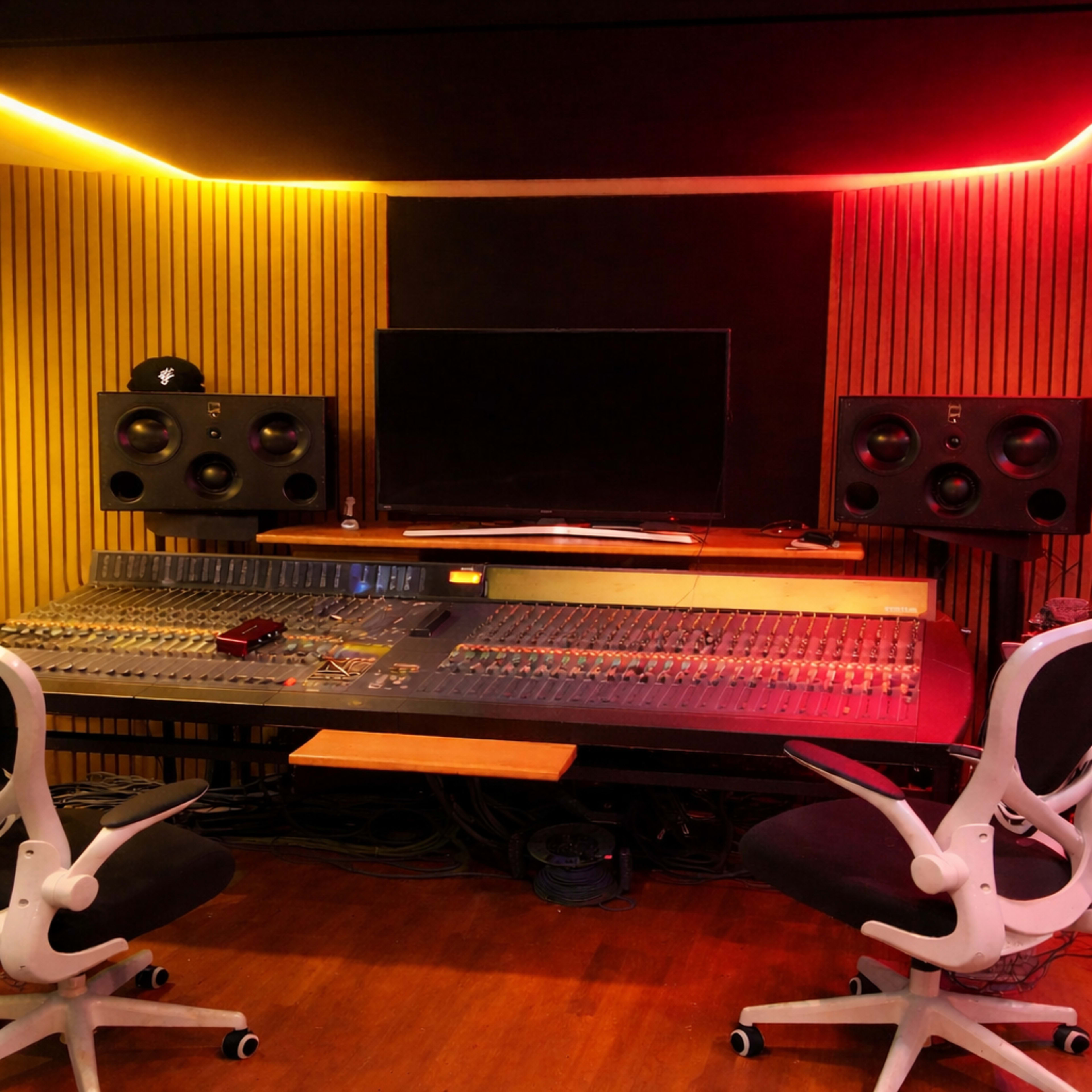 The image shows a music studio with a large mixing console, two studio monitors, a television screen, and two chairs.