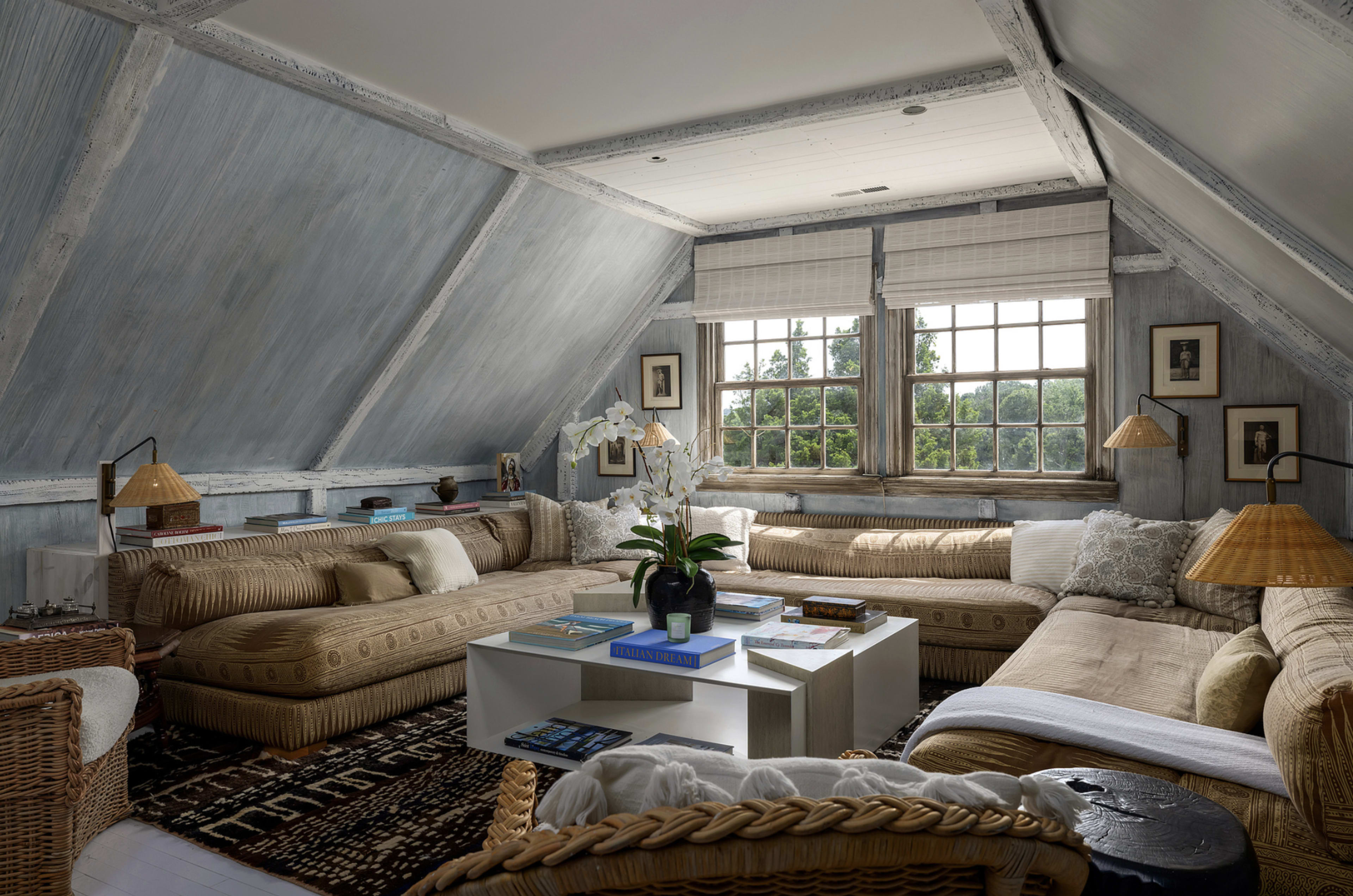 The image shows a cozy attic living room featuring large sectional sofas, a central coffee table, and windows that provide a view of the outdoors.