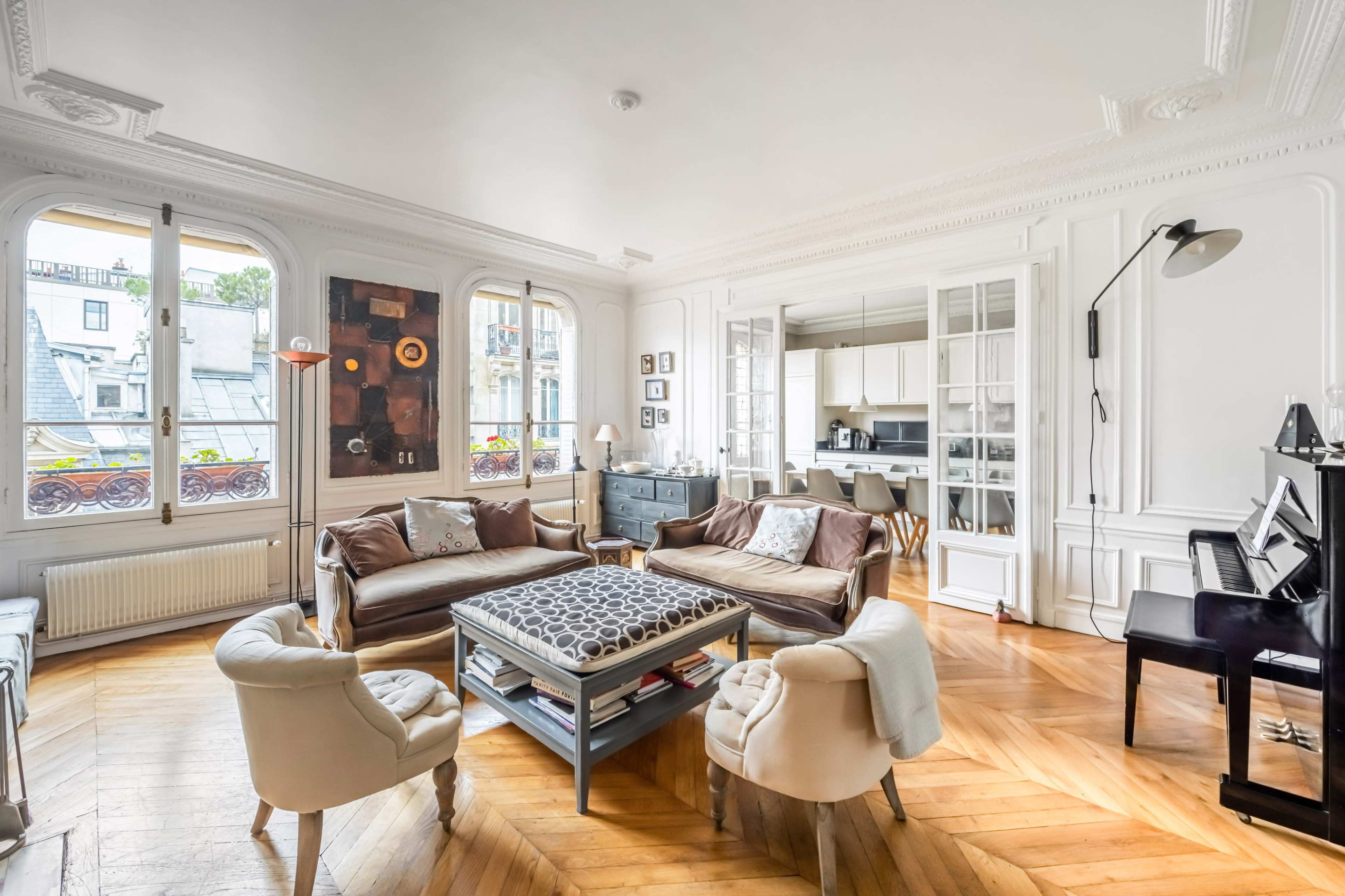 The image shows a well-lit living room with elegant furnishings, large windows, and wooden flooring.