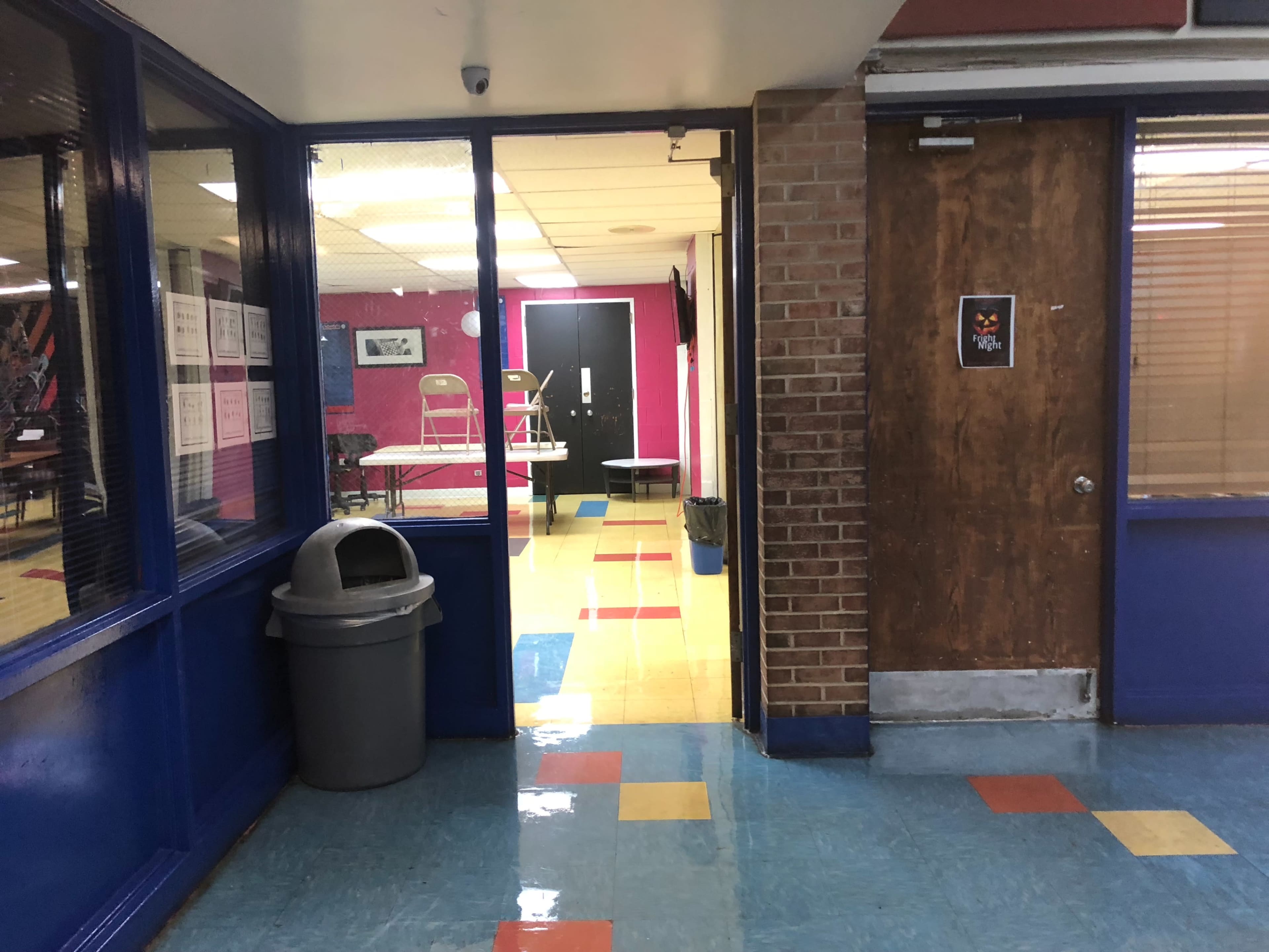 A hallway with a trash can, leading to a brightly colored room with chairs and tables.