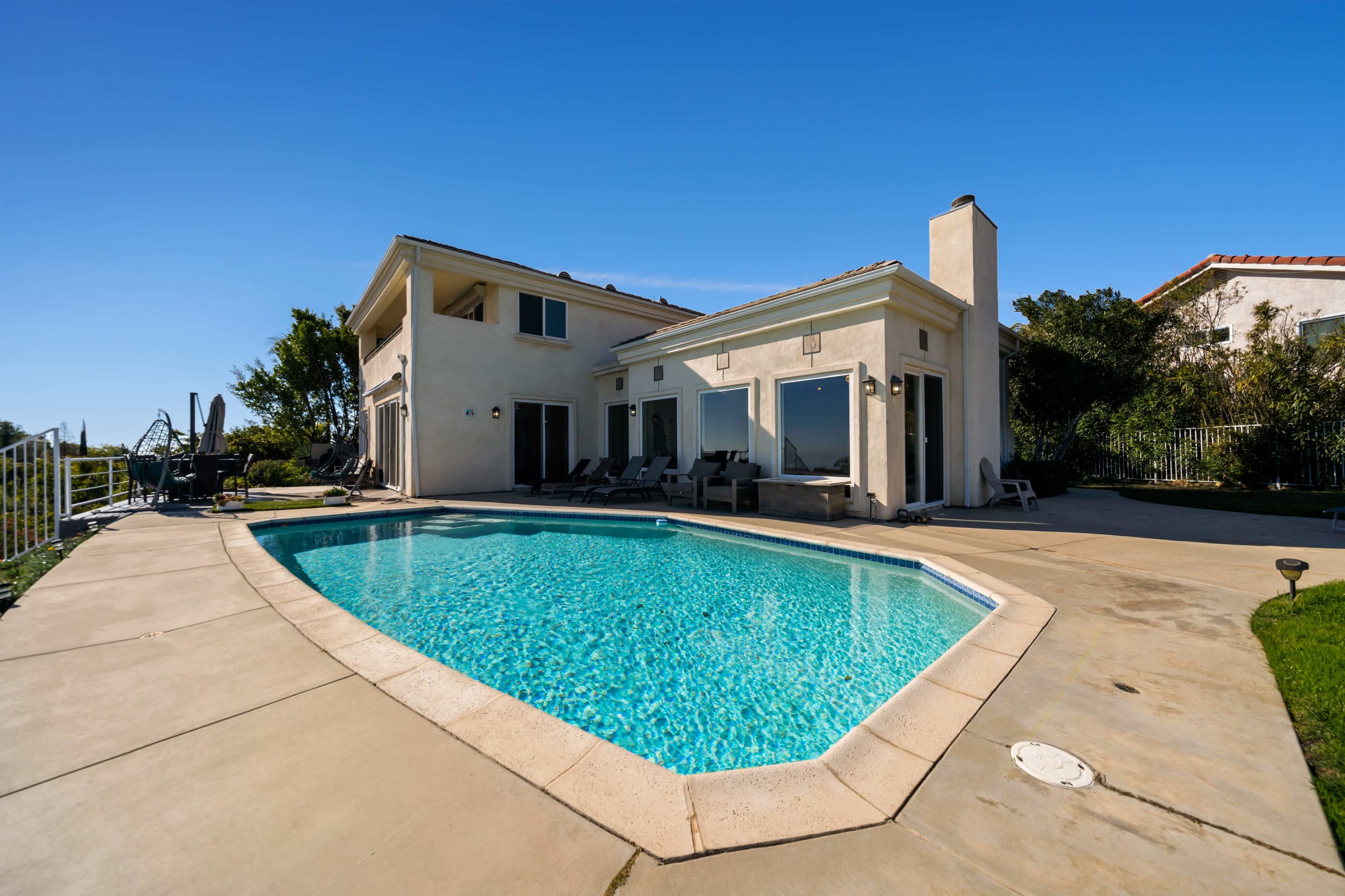 The image shows a home with a swimming pool surrounded by a concrete patio and green landscaping under a clear blue sky.