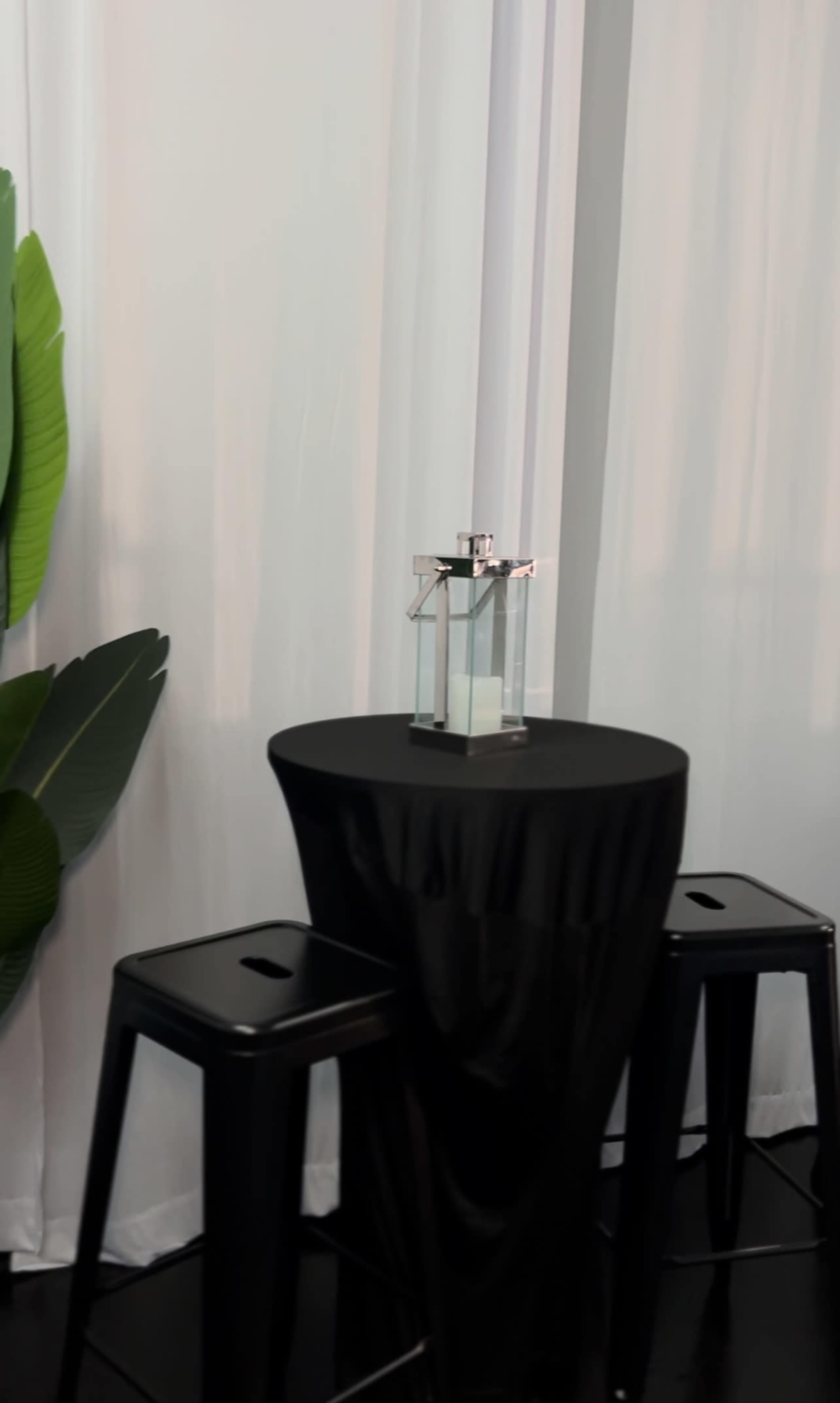 A clear perfume bottle is placed on a small black round table surrounded by two black stools, with large green leaves in the background.