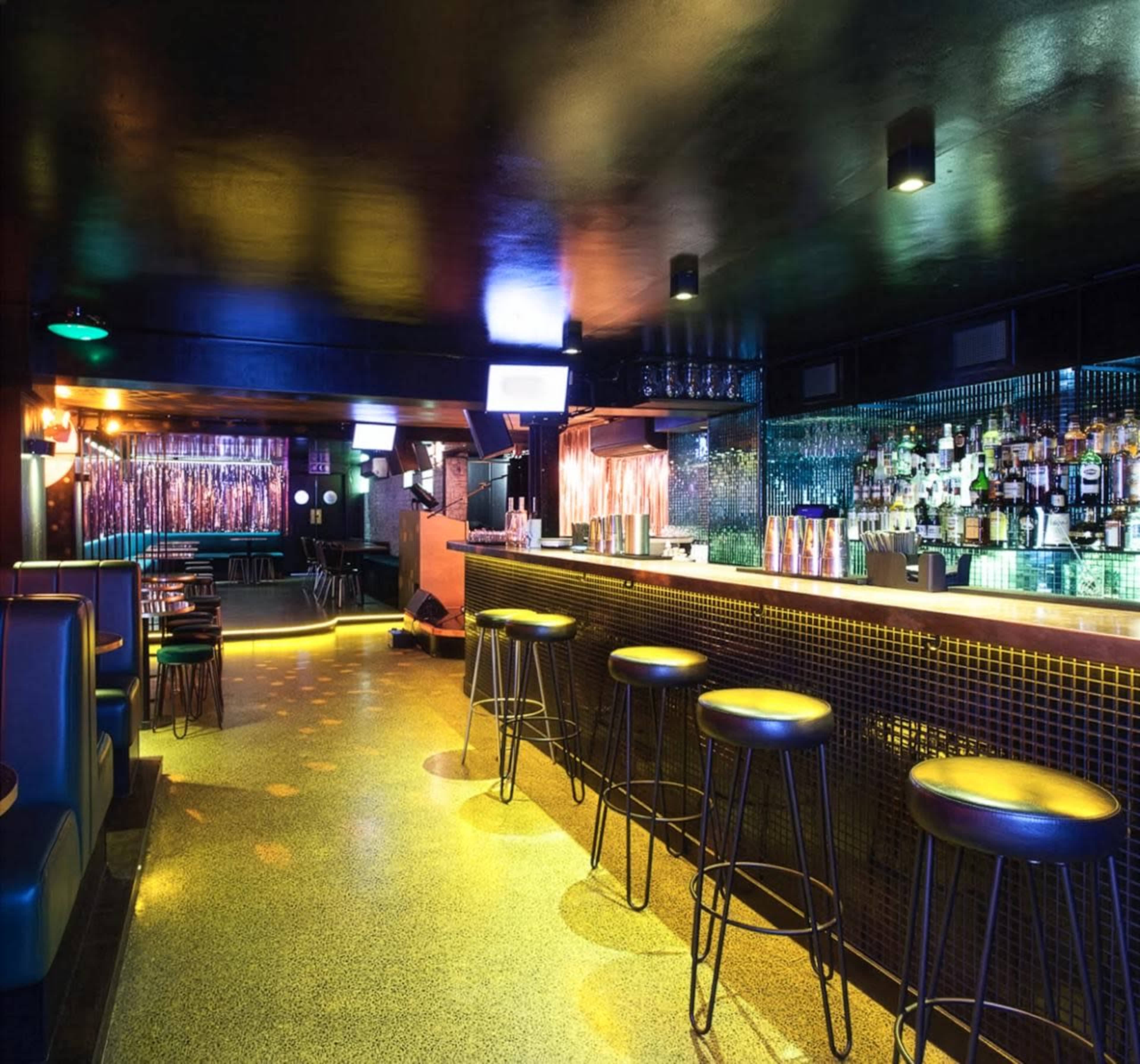 The image shows a dimly lit bar area with a polished counter, high stools, and a colorful backdrop adorned with bottles and lights.