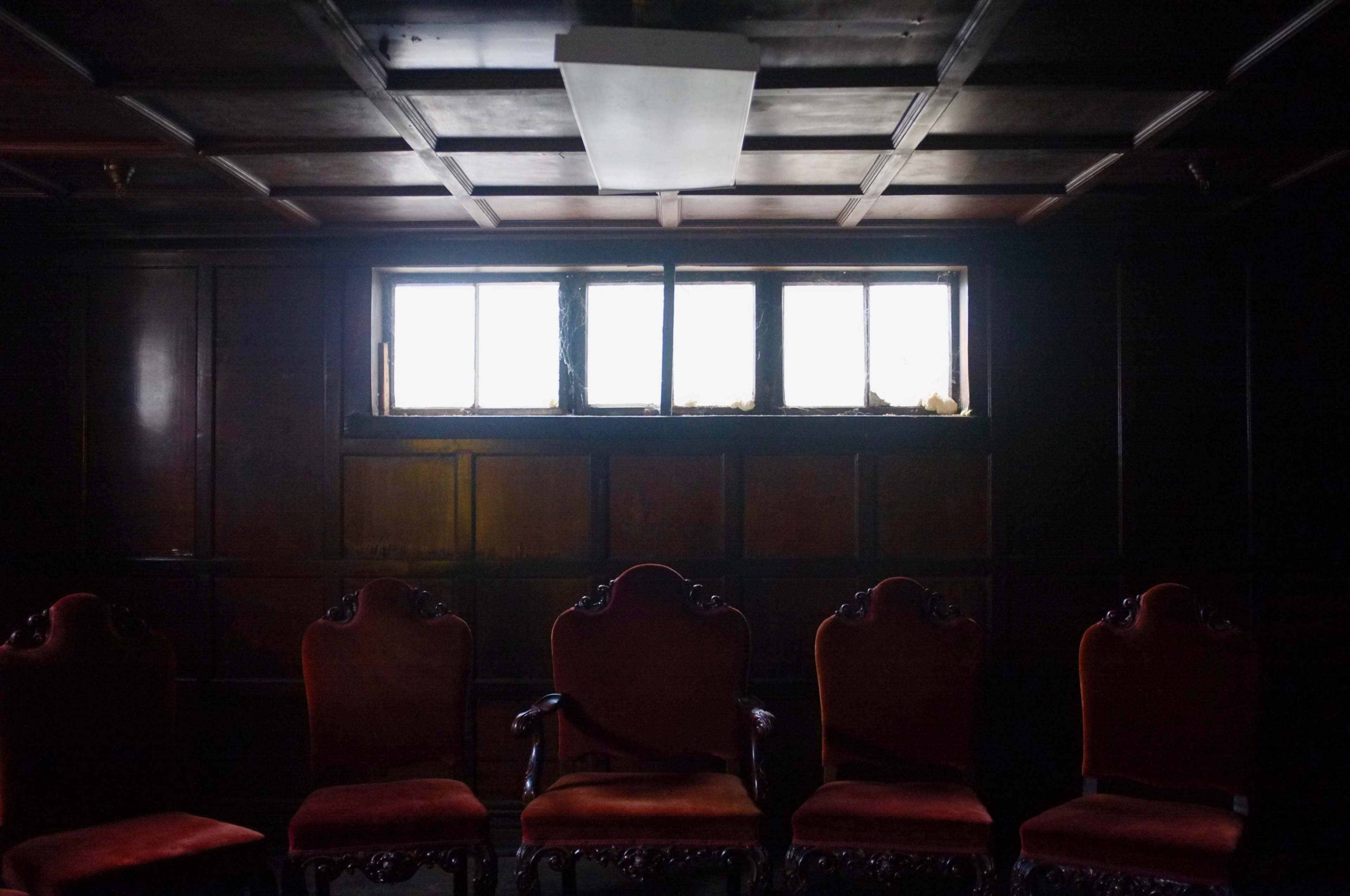 The image shows a dimly lit room with a row of ornate, upholstered chairs facing a small window.