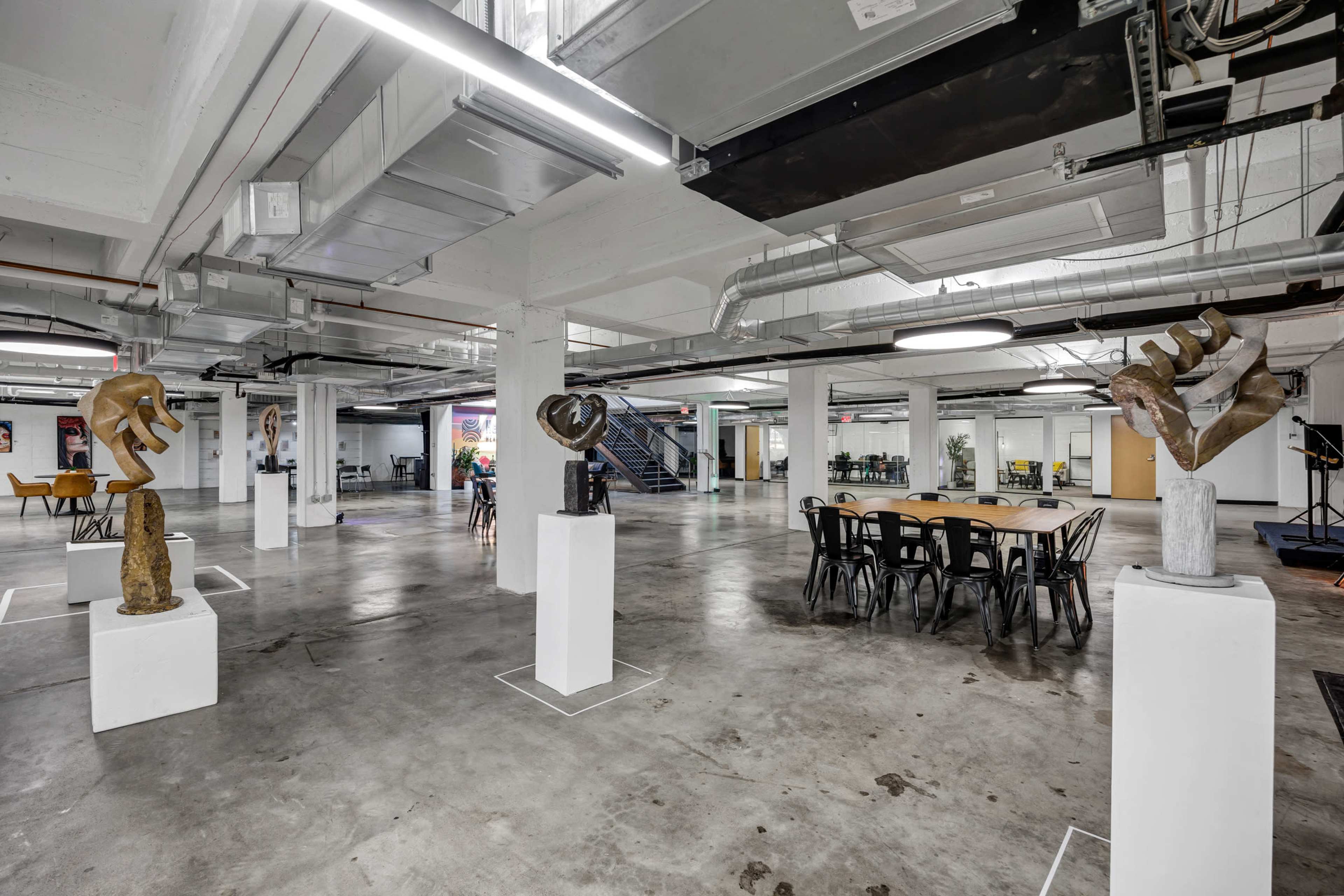 The image shows an industrial-style interior space with concrete flooring, featuring various sculptures displayed on white pedestals and a large communal table surrounded by black chairs.