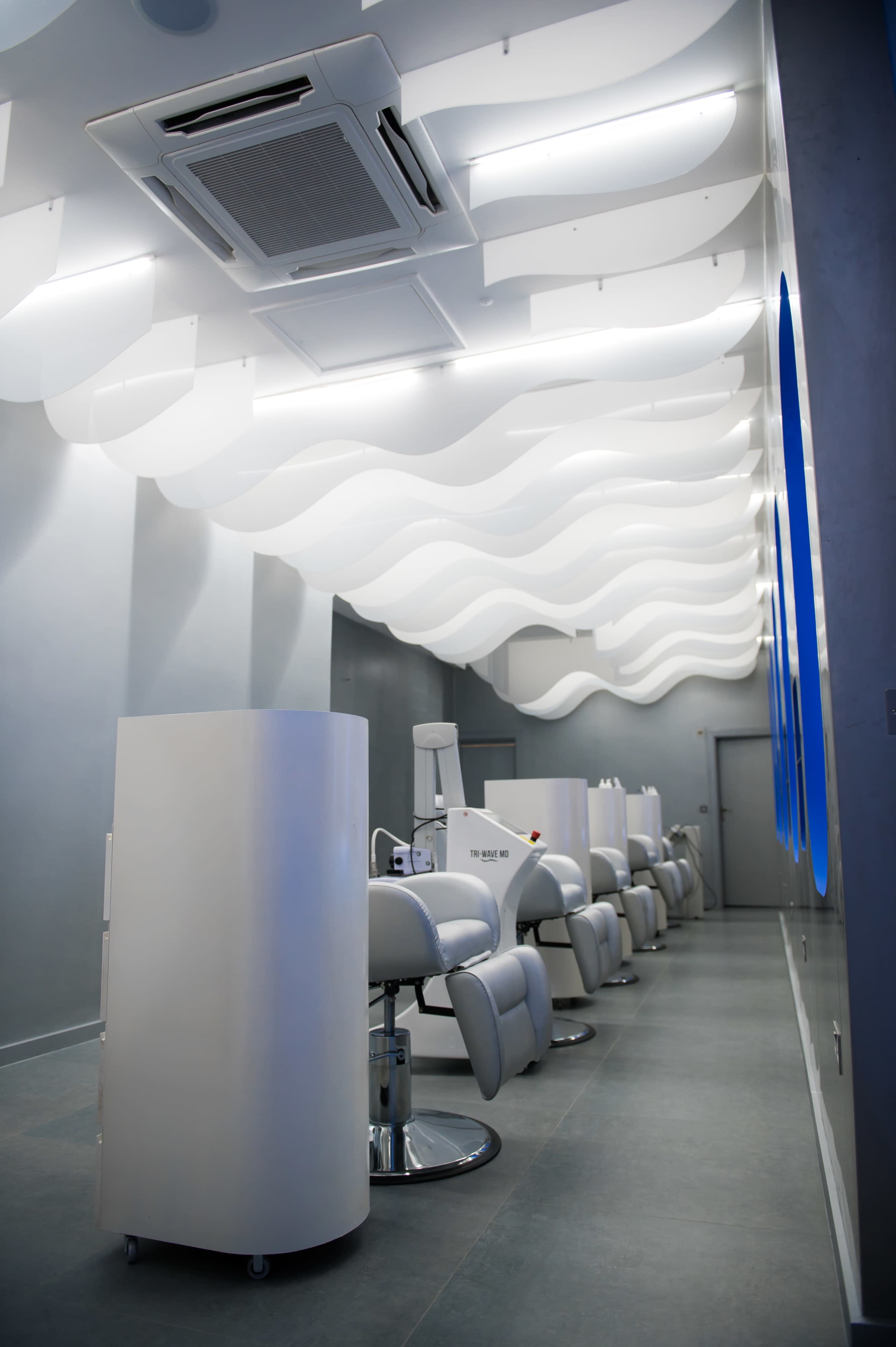 A modern salon interior with white, wave-shaped ceiling panels and several styling stations lined up along the wall.