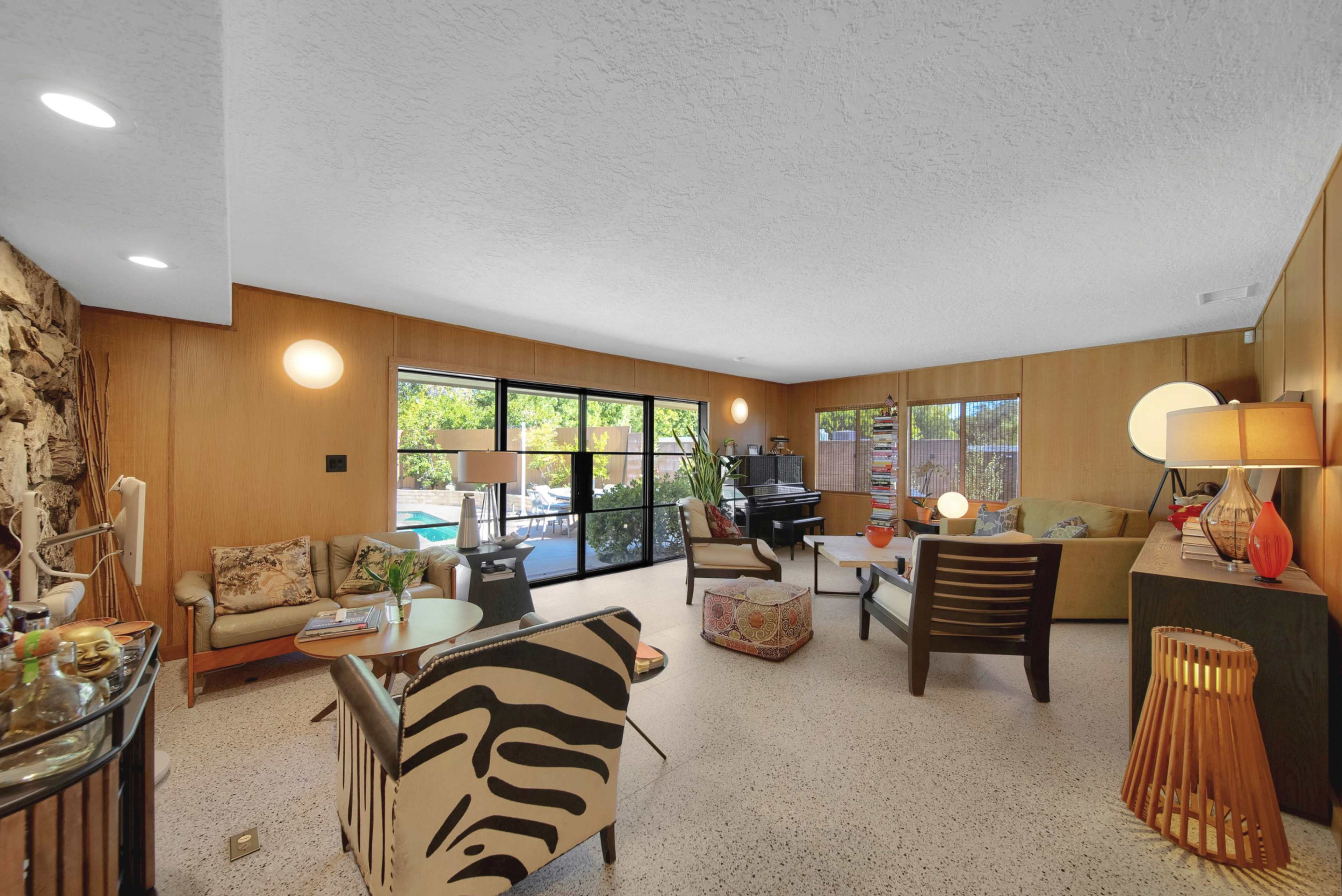 The image depicts a mid-century modern living room featuring large windows, patterned furniture, and a piano in the background.