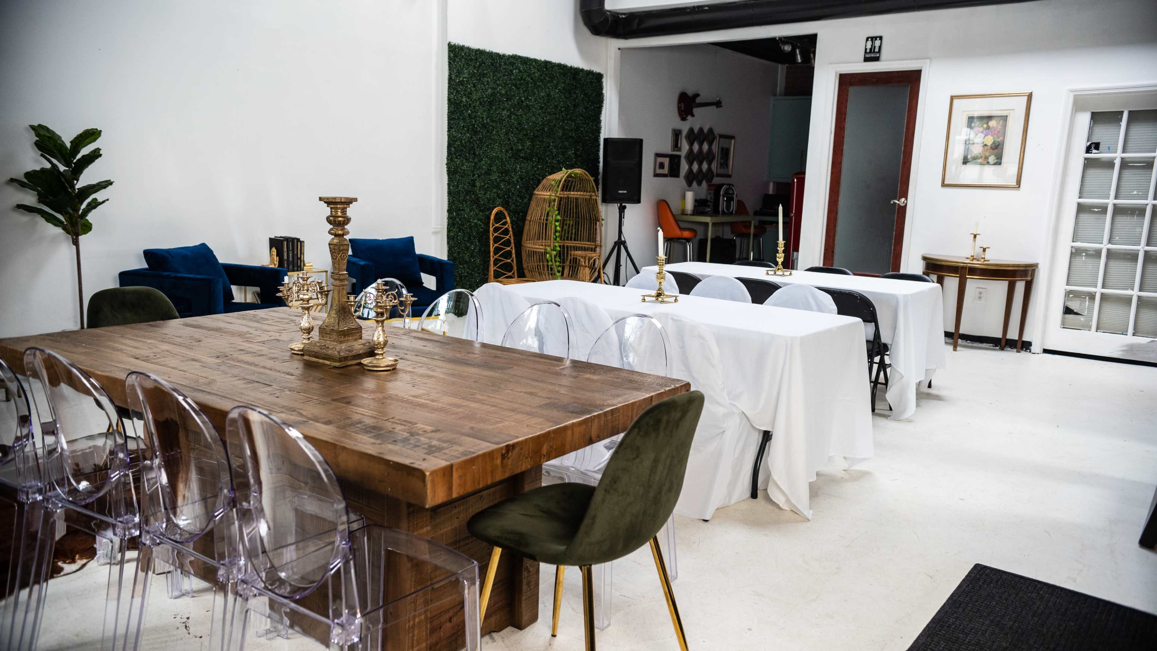 The image shows a modern event space featuring a large wooden table surrounded by transparent chairs and several covered tables in a well-lit room with greenery decorations.