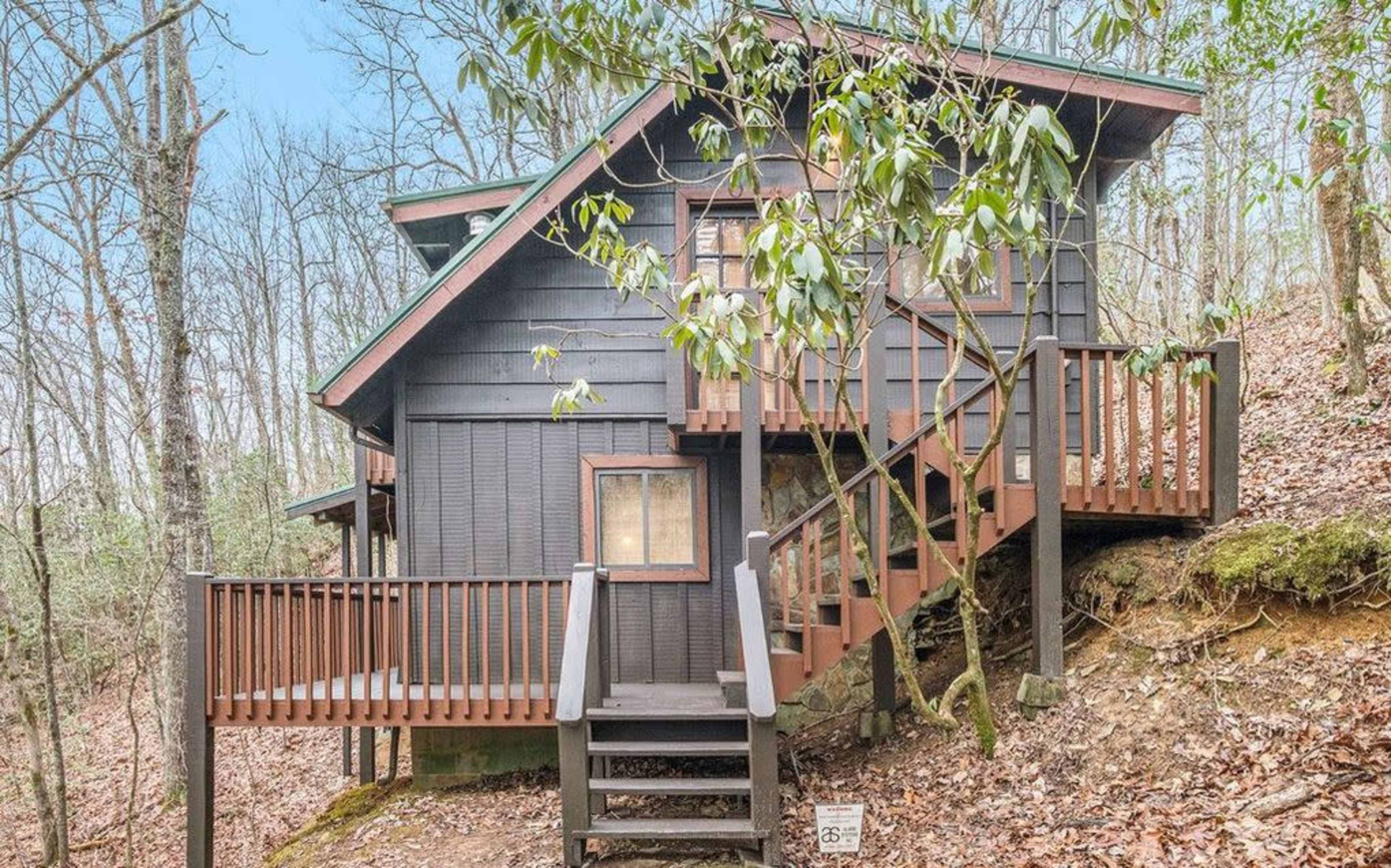 A dark-stained, two-story cabin is nestled among trees on a sloped lot, featuring a staircase leading to its entrance.