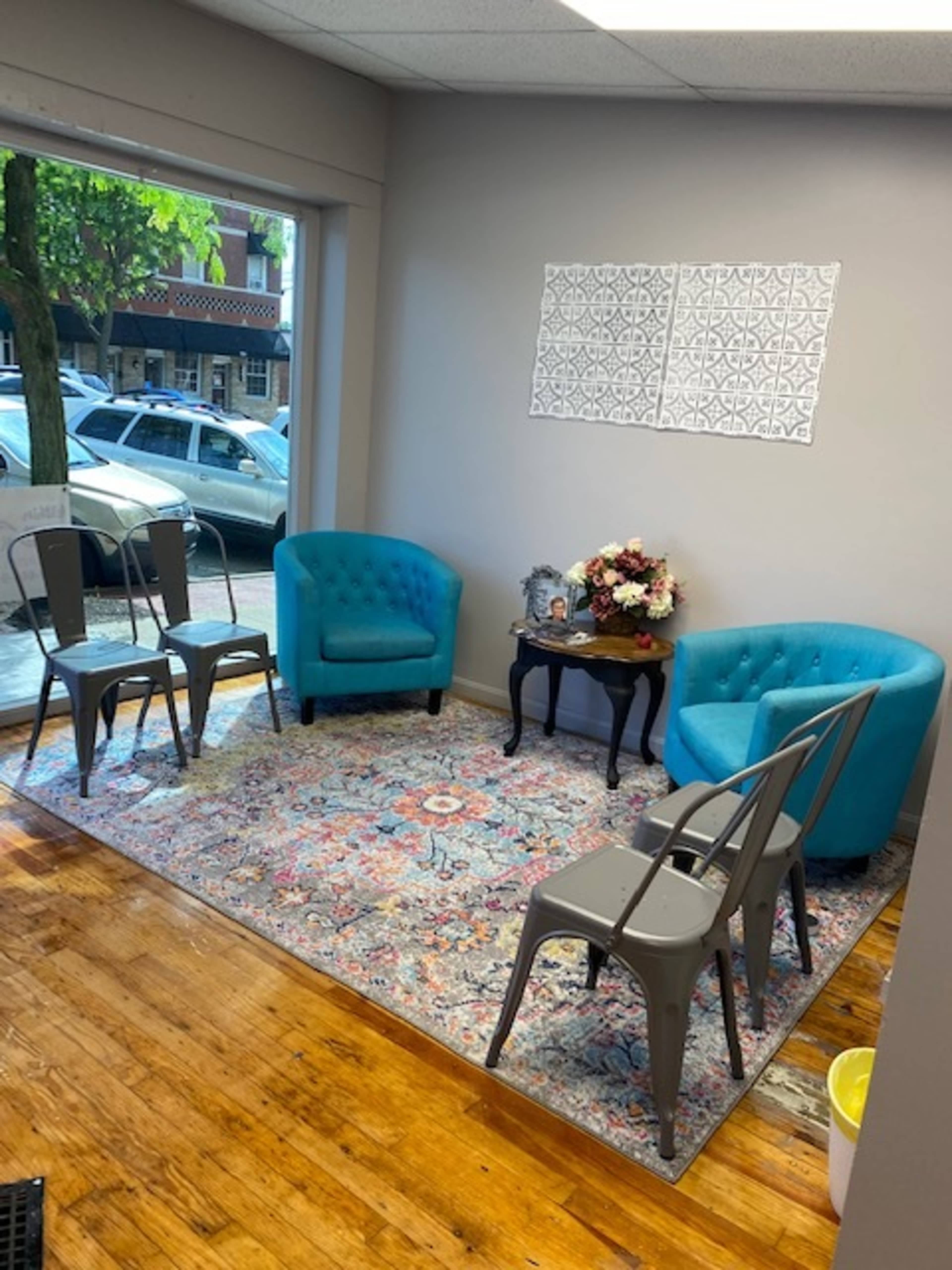 The image shows a small waiting area with two turquoise chairs, a small table, a floral arrangement, and three metal chairs, all set on a colorful area rug.