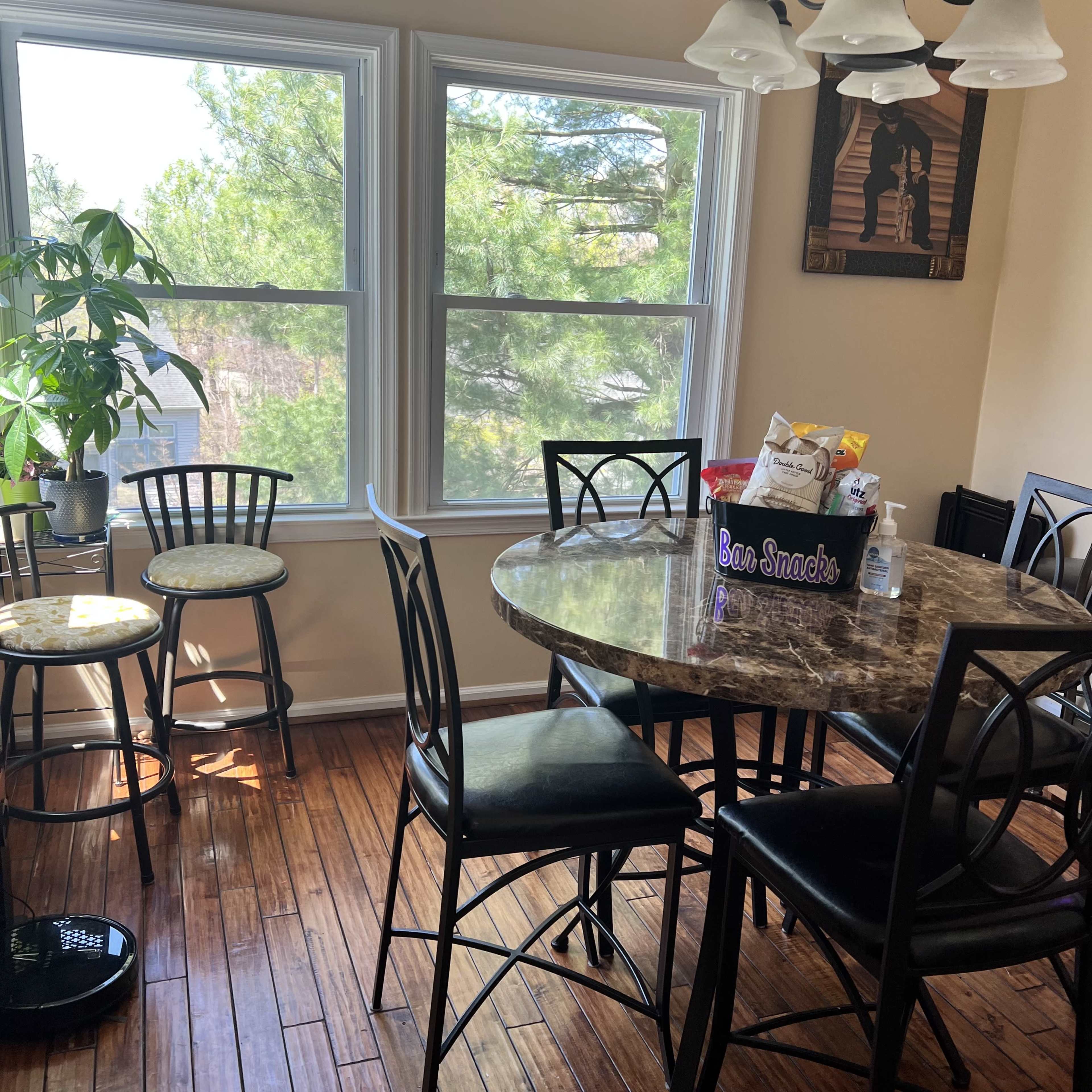 A round marble dining table with black chairs sits in a brightly lit room, featuring large windows that overlook greenery outside.