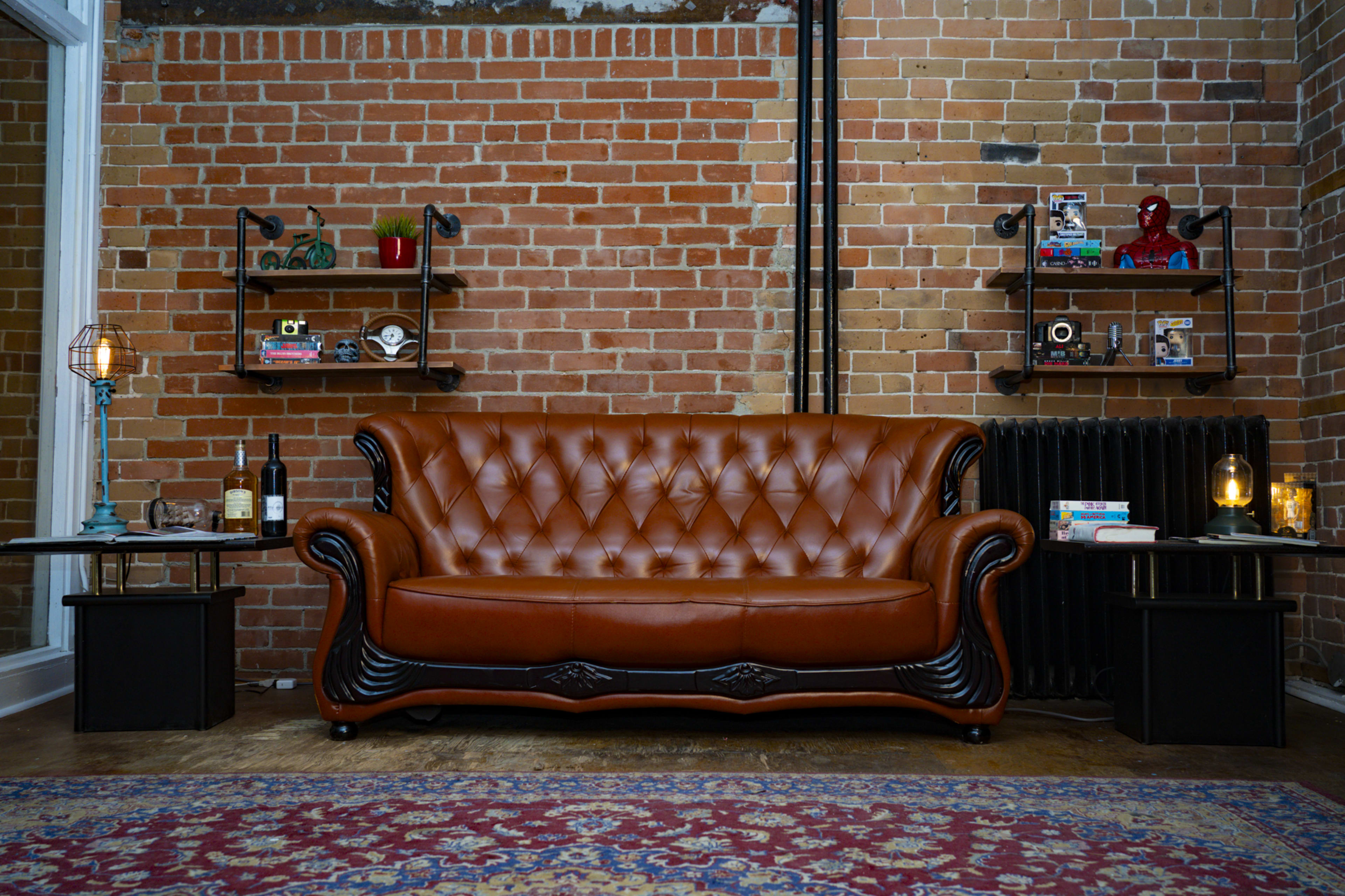 The image features a brown tufted sofa placed against a brick wall, with two shelves displaying various items on either side and a small table with a lamp beside the sofa.