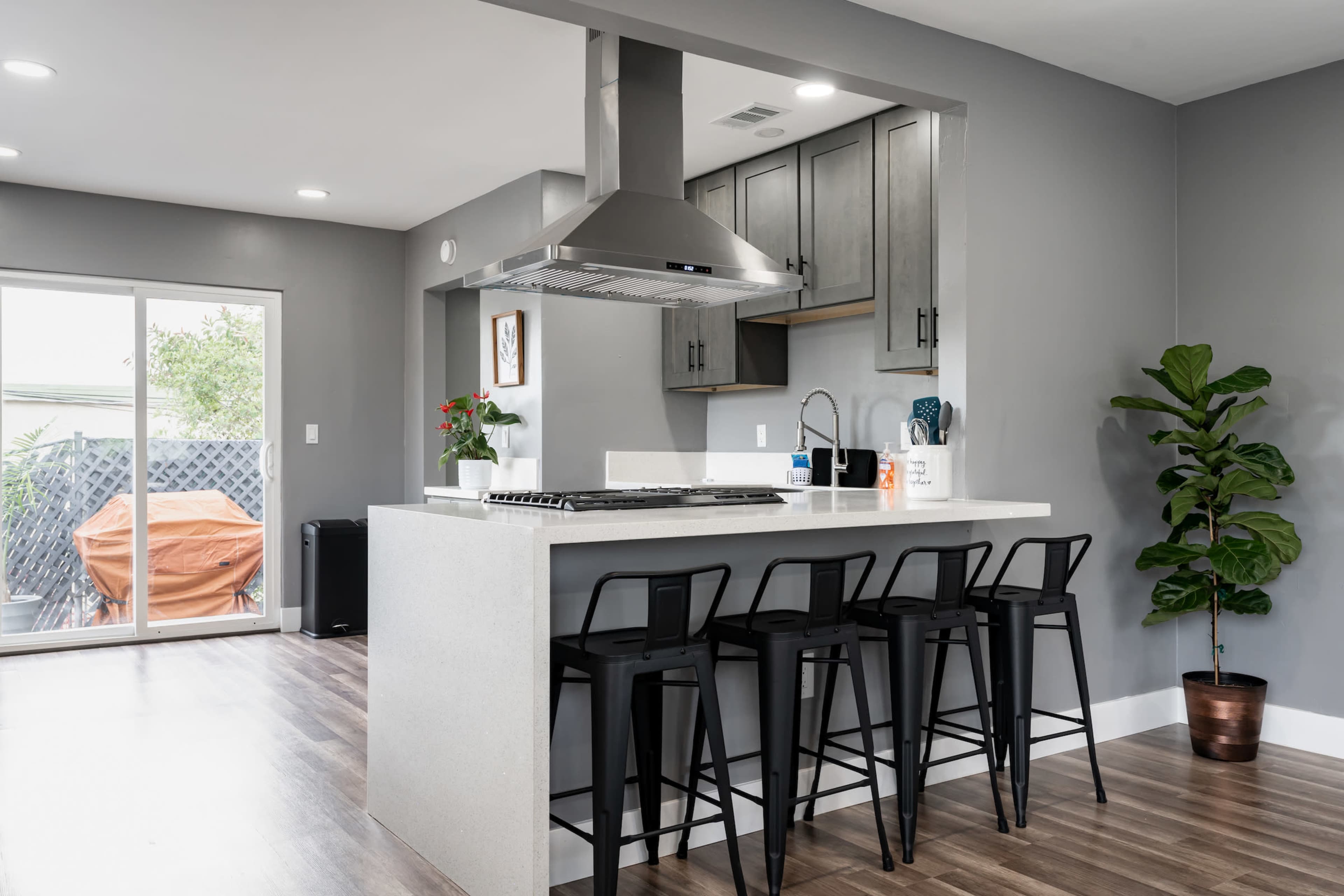 The image shows a modern kitchen with a central island, four black bar stools, stainless steel appliances, and a large sliding door leading to an outdoor area.