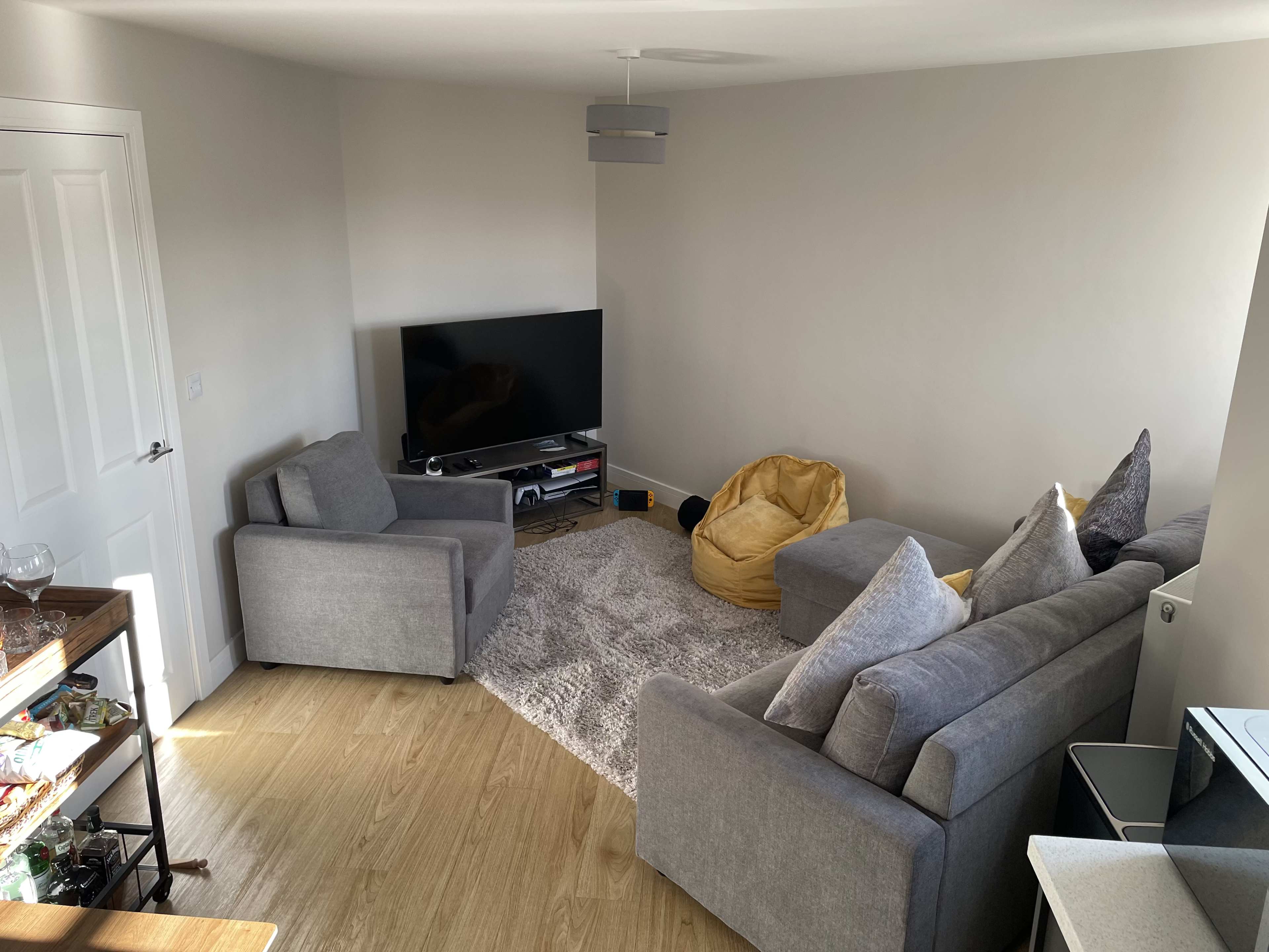 The image shows a modern living room with two gray armchairs, a beige sofa with decorative pillows, a television on a stand, and a yellow bean bag on a light gray area rug.