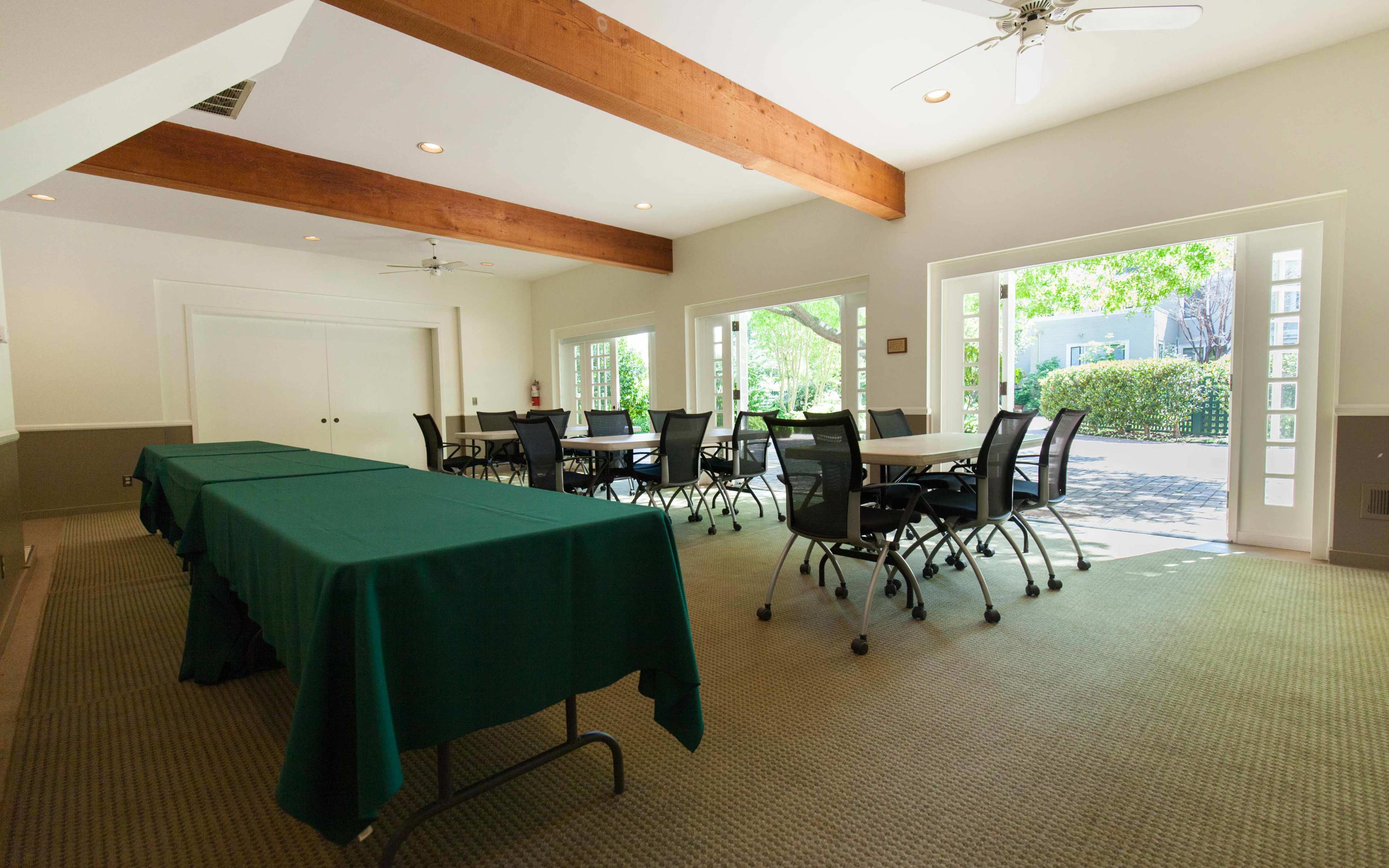 A well-lit meeting room featuring tables covered with green cloths and rows of black chairs, surrounded by large windows overlooking a garden.