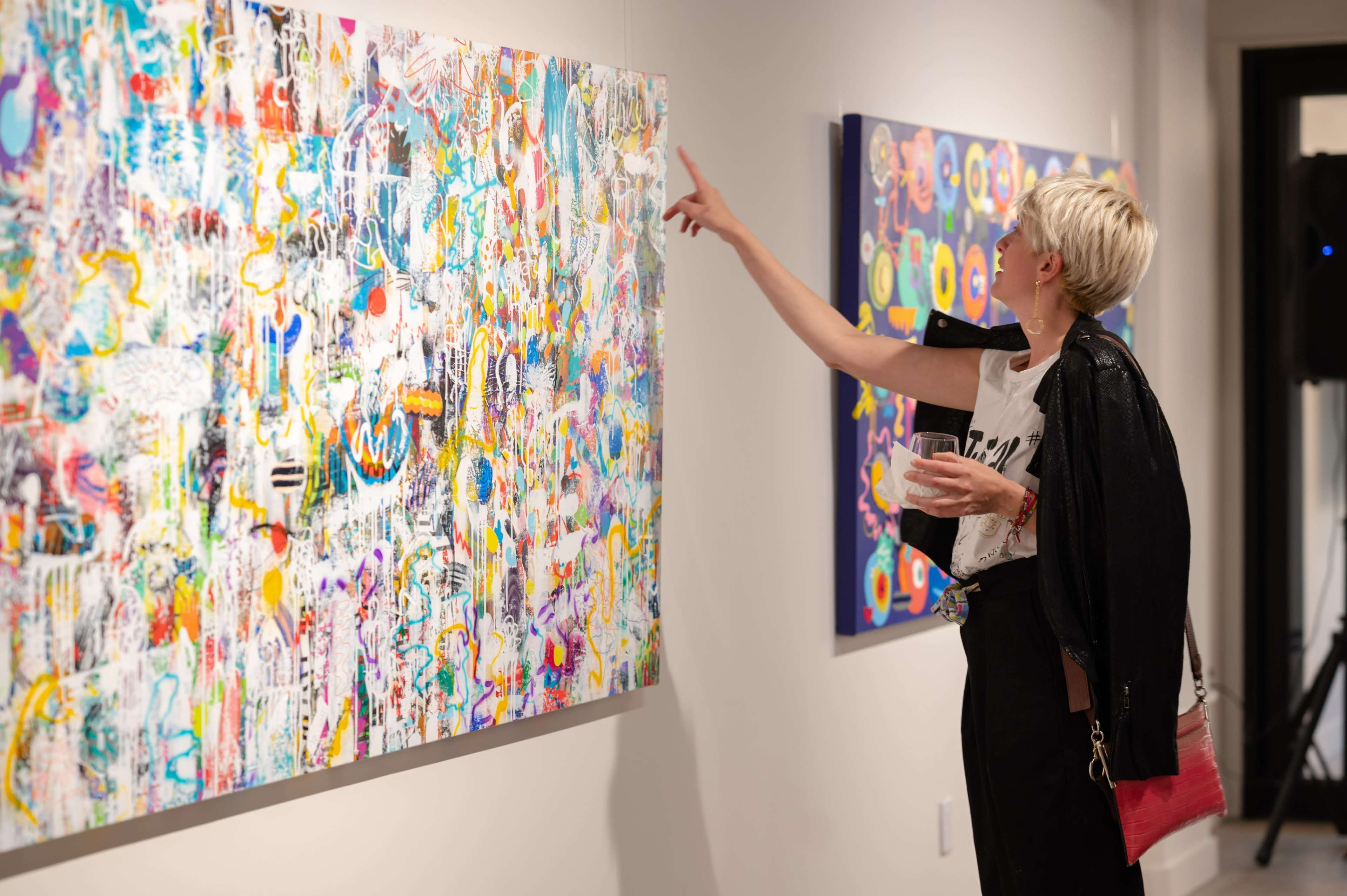 A woman holds a drink while pointing at a colorful abstract painting in an art gallery.