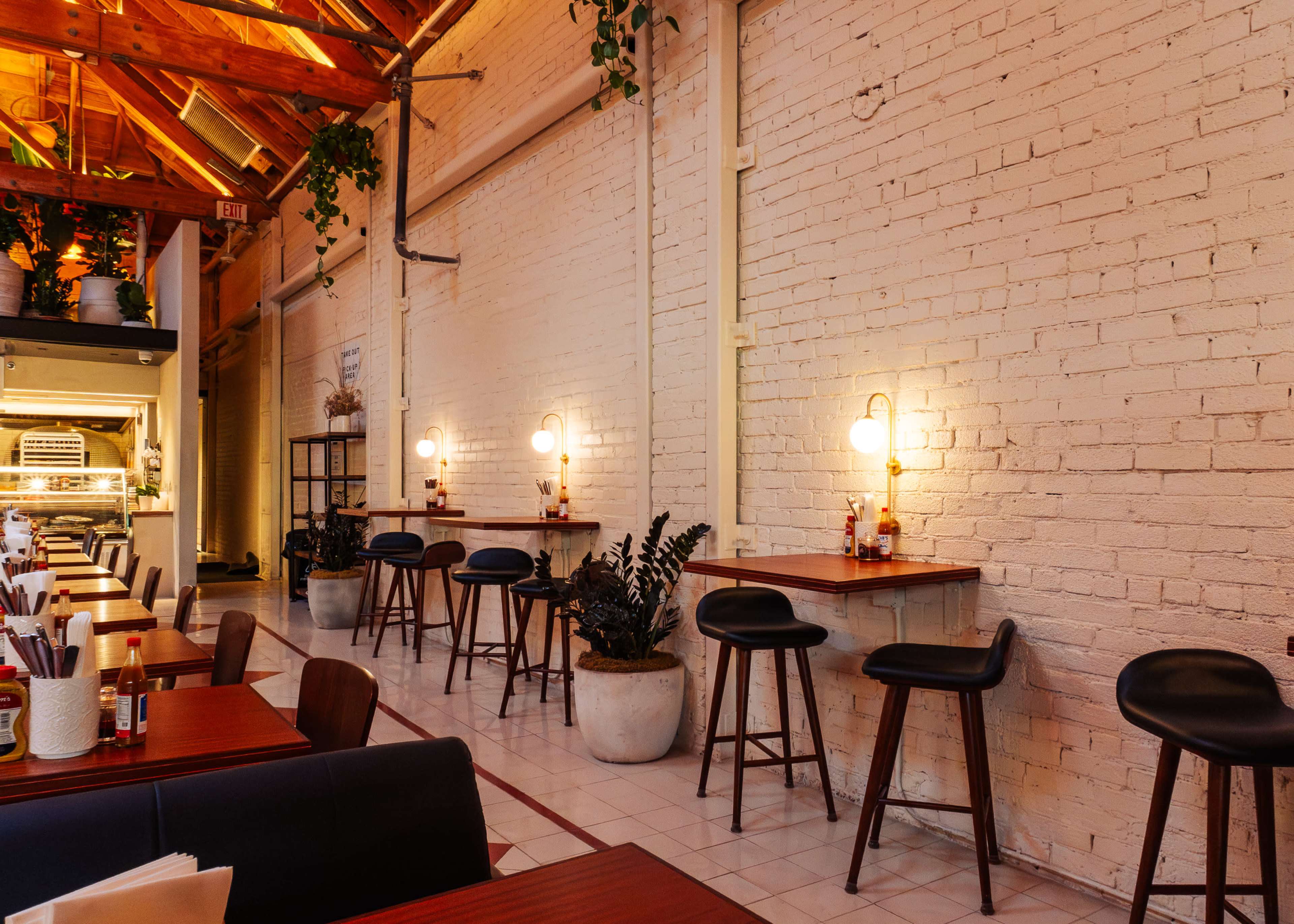 The image shows a modern restaurant interior with white brick walls, wooden beams, and small tables against the wall, each paired with high stools and decorative plants.