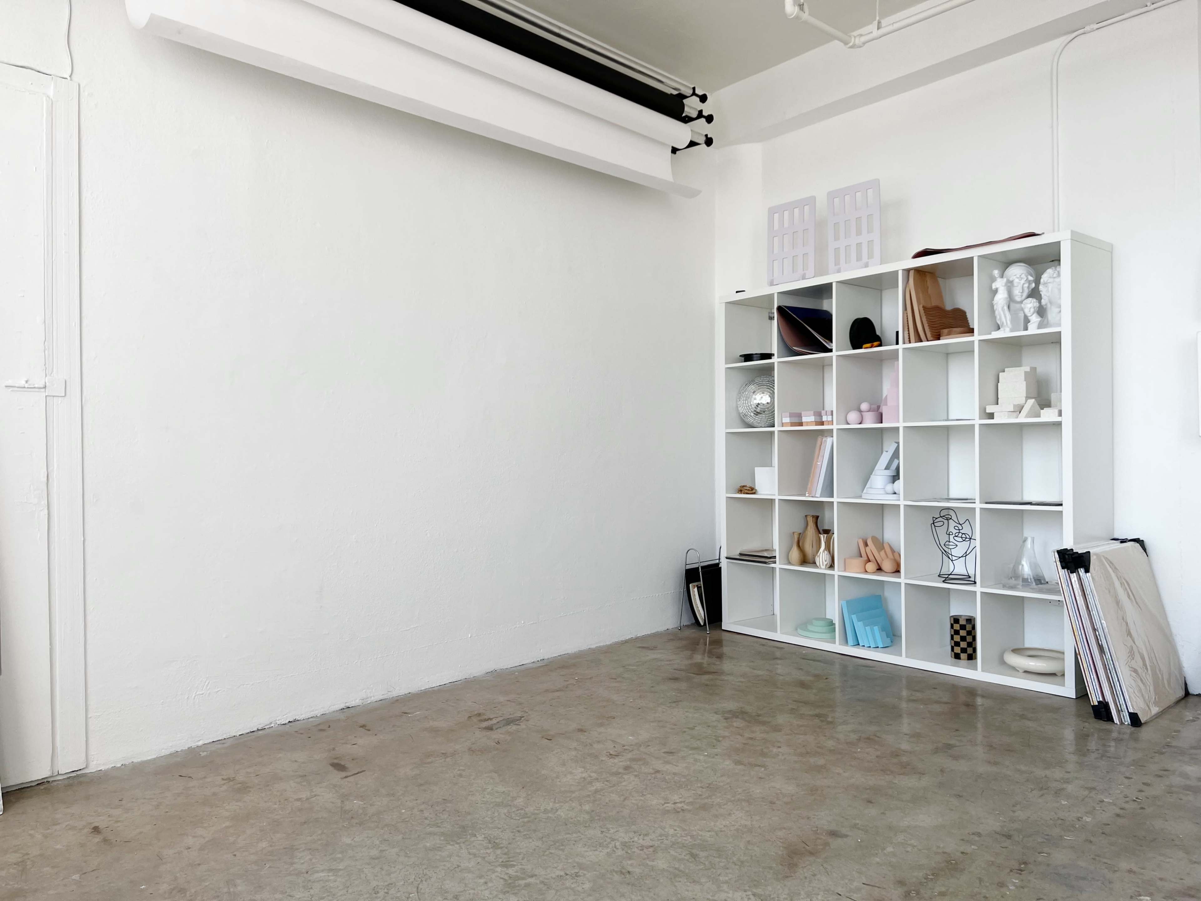 A minimalist studio features a white bookshelf filled with various art objects and sculptures against a plain wall, with a polished concrete floor.