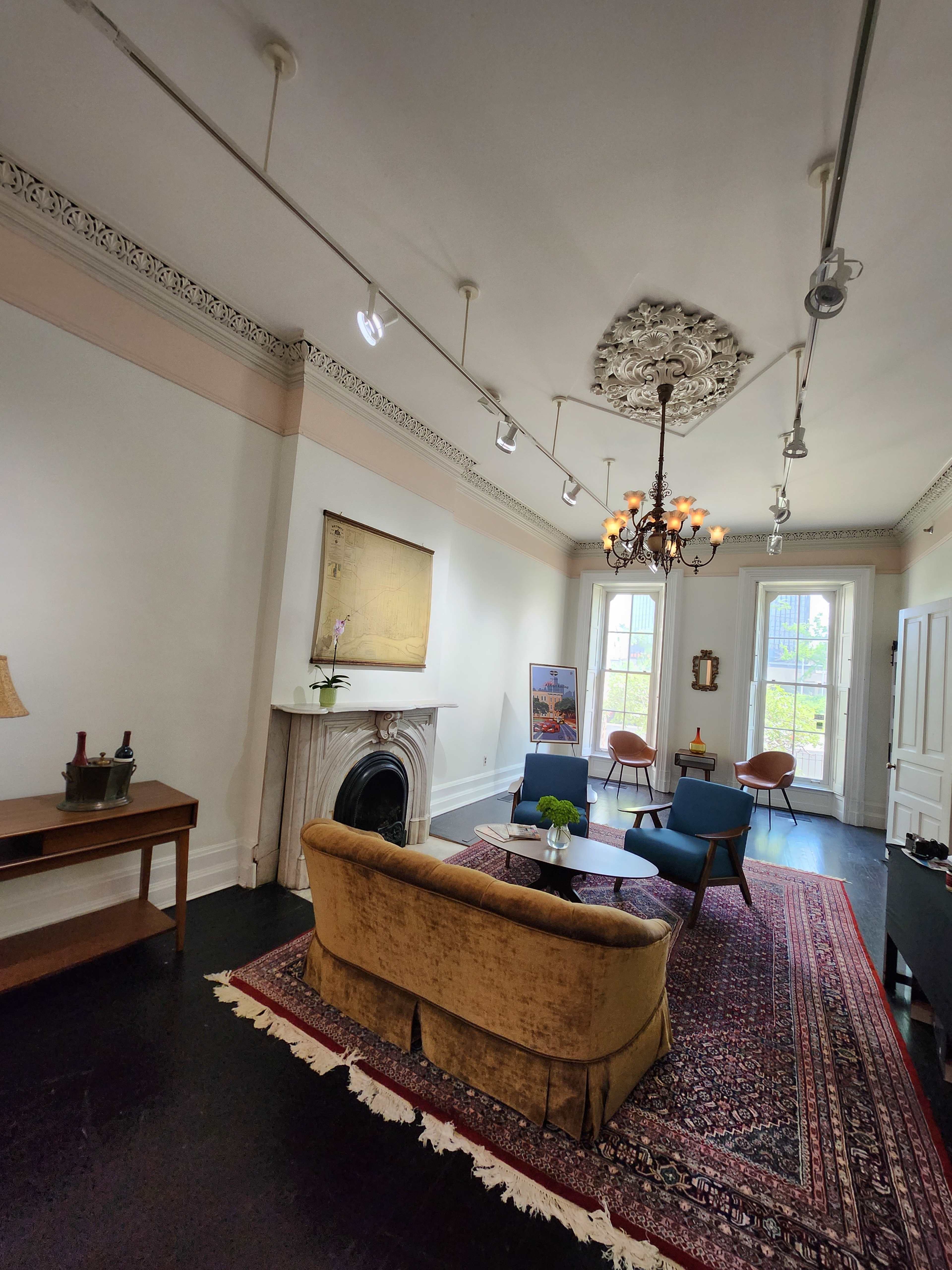 A spacious room with a chandelier, a vintage sofa, a coffee table, and a large window, featuring a decorative fireplace and ornate ceiling trim.