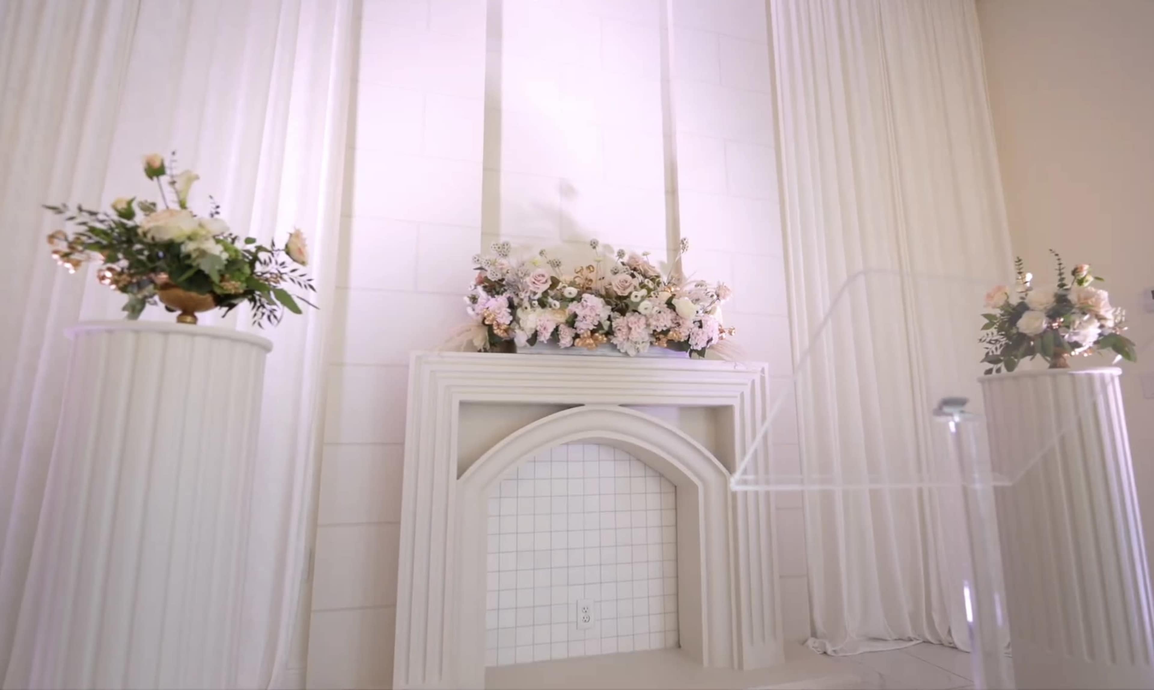 The image shows an elegantly decorated altar with a floral arrangement on a white mantel, flanked by two columns with smaller floral arrangements, set against a light-colored backdrop.