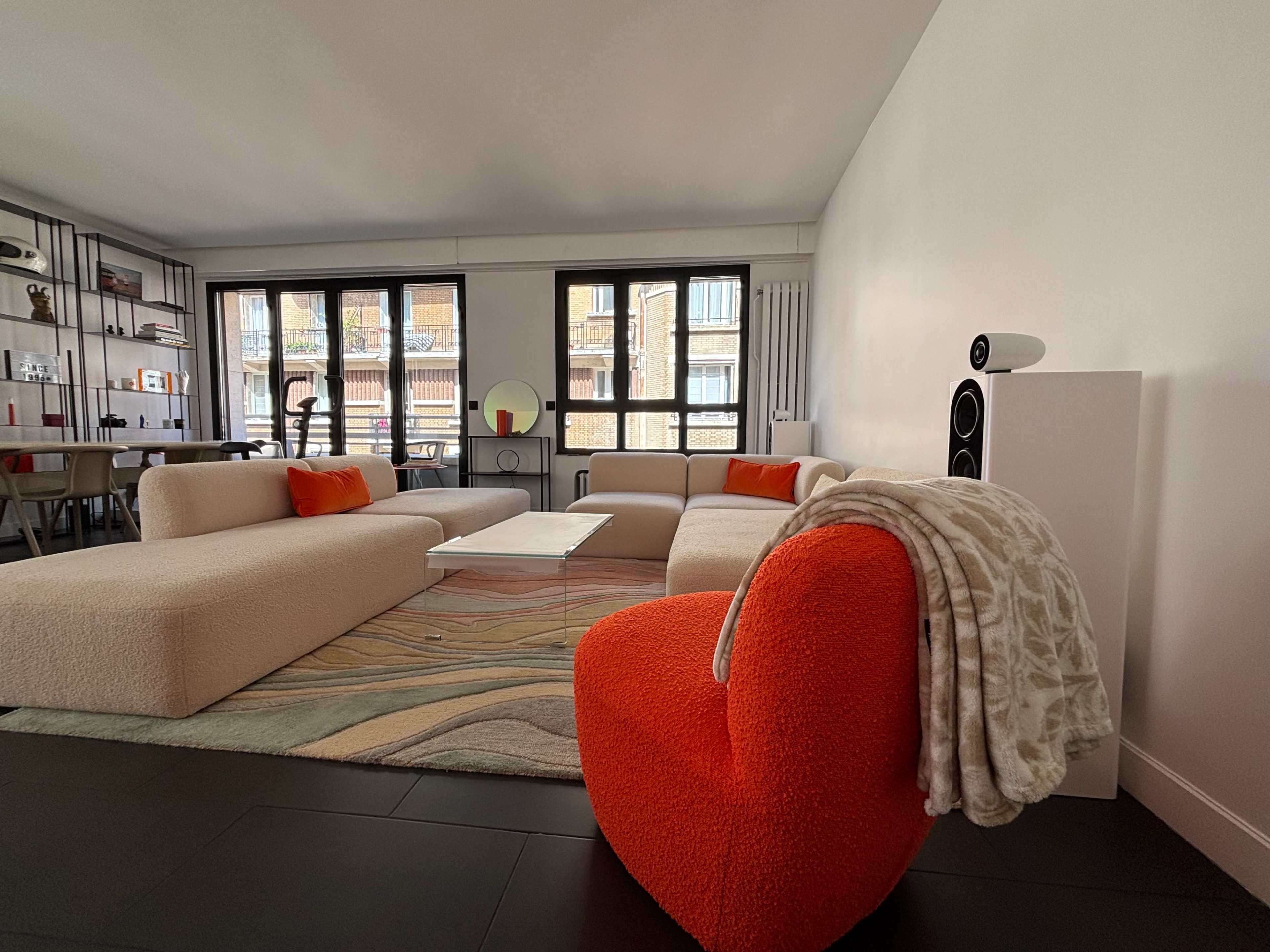 A modern living room features a light-colored sofa, an orange accent chair, a white coffee table, and large windows overlooking a cityscape.