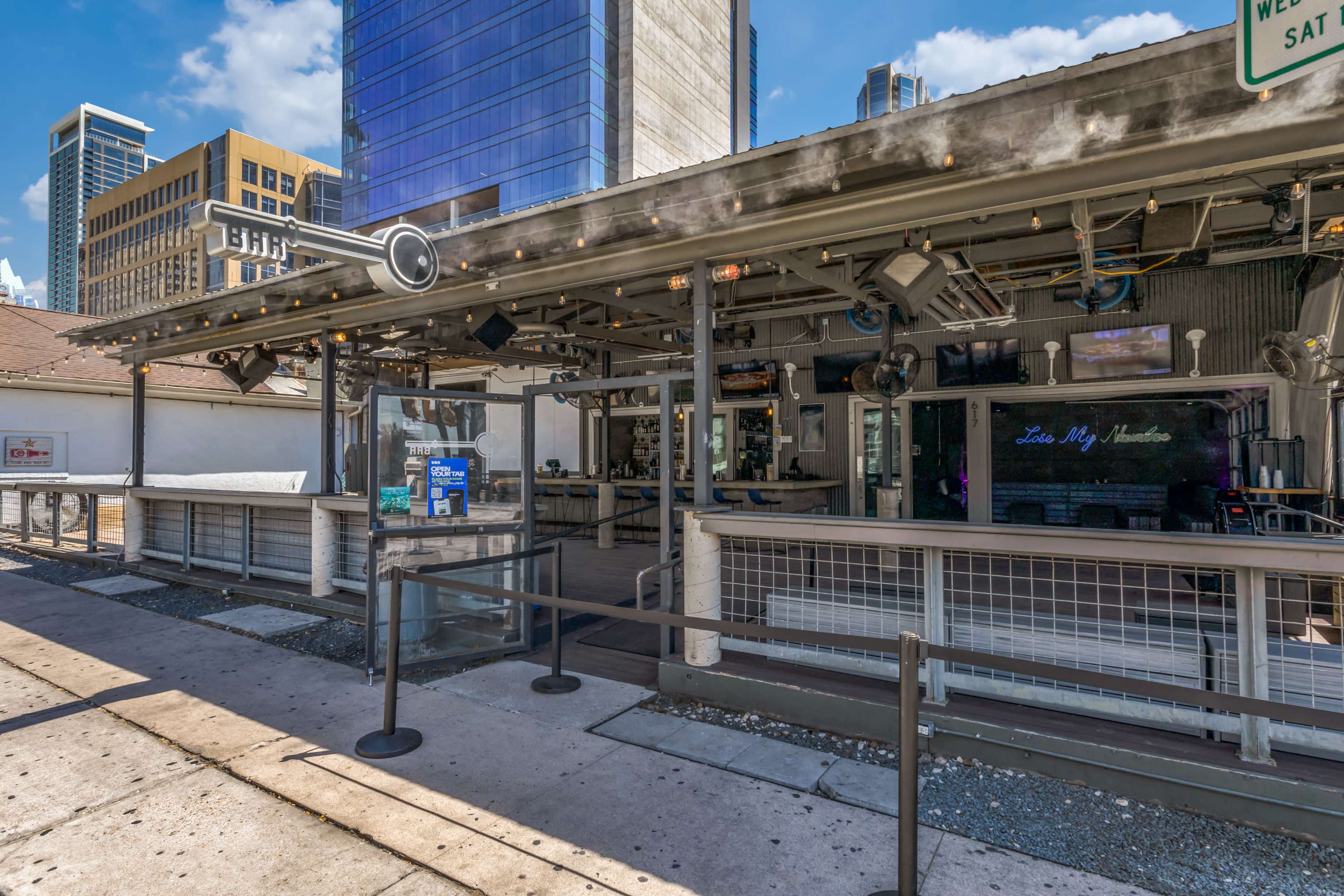 The image shows an outdoor bar with a covered seating area, surrounded by modern buildings in a city setting.