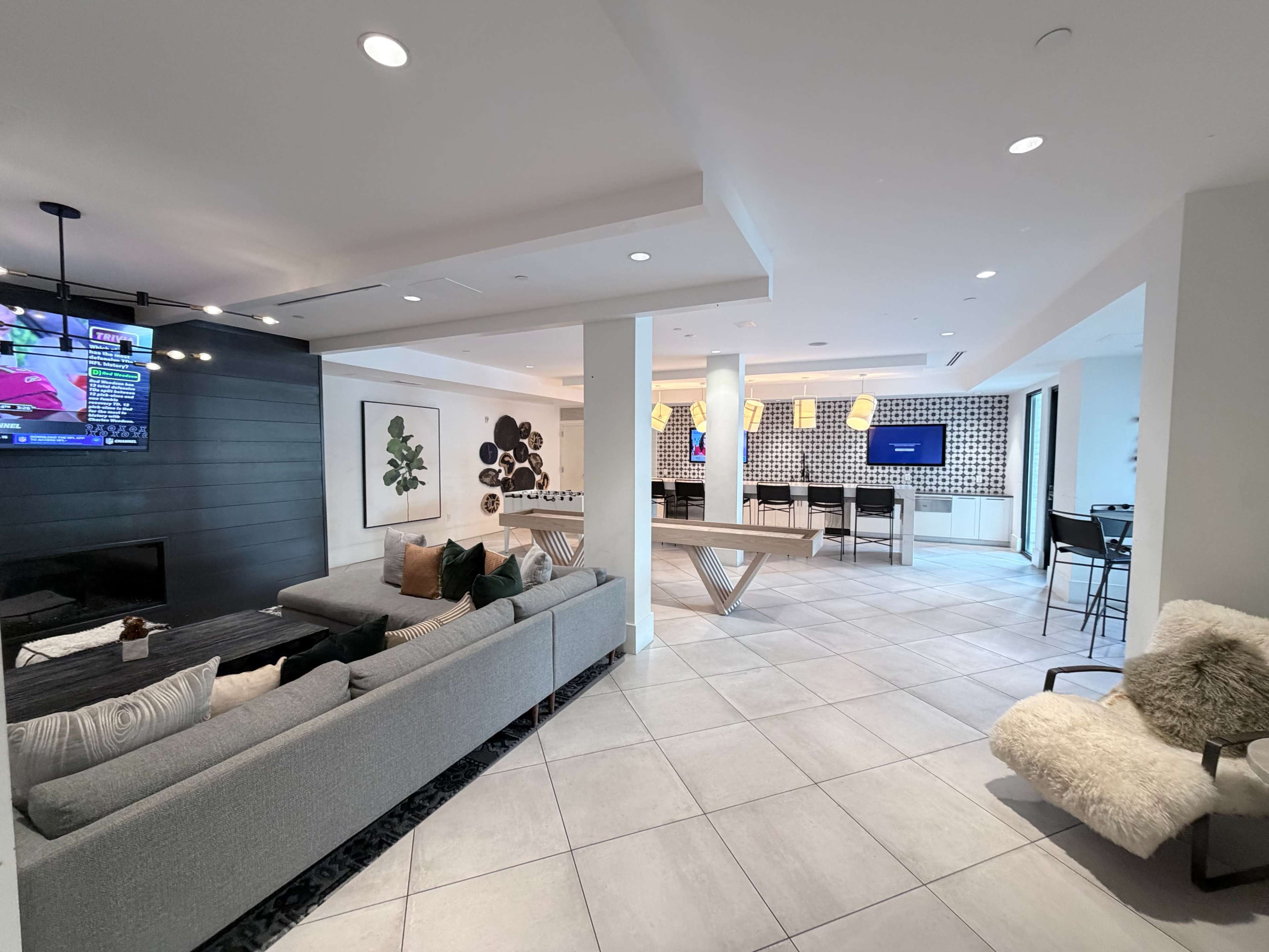 The image shows a modern open-concept living area featuring a sectional sofa, a wall-mounted television, and a dining area with high stools and a large table.