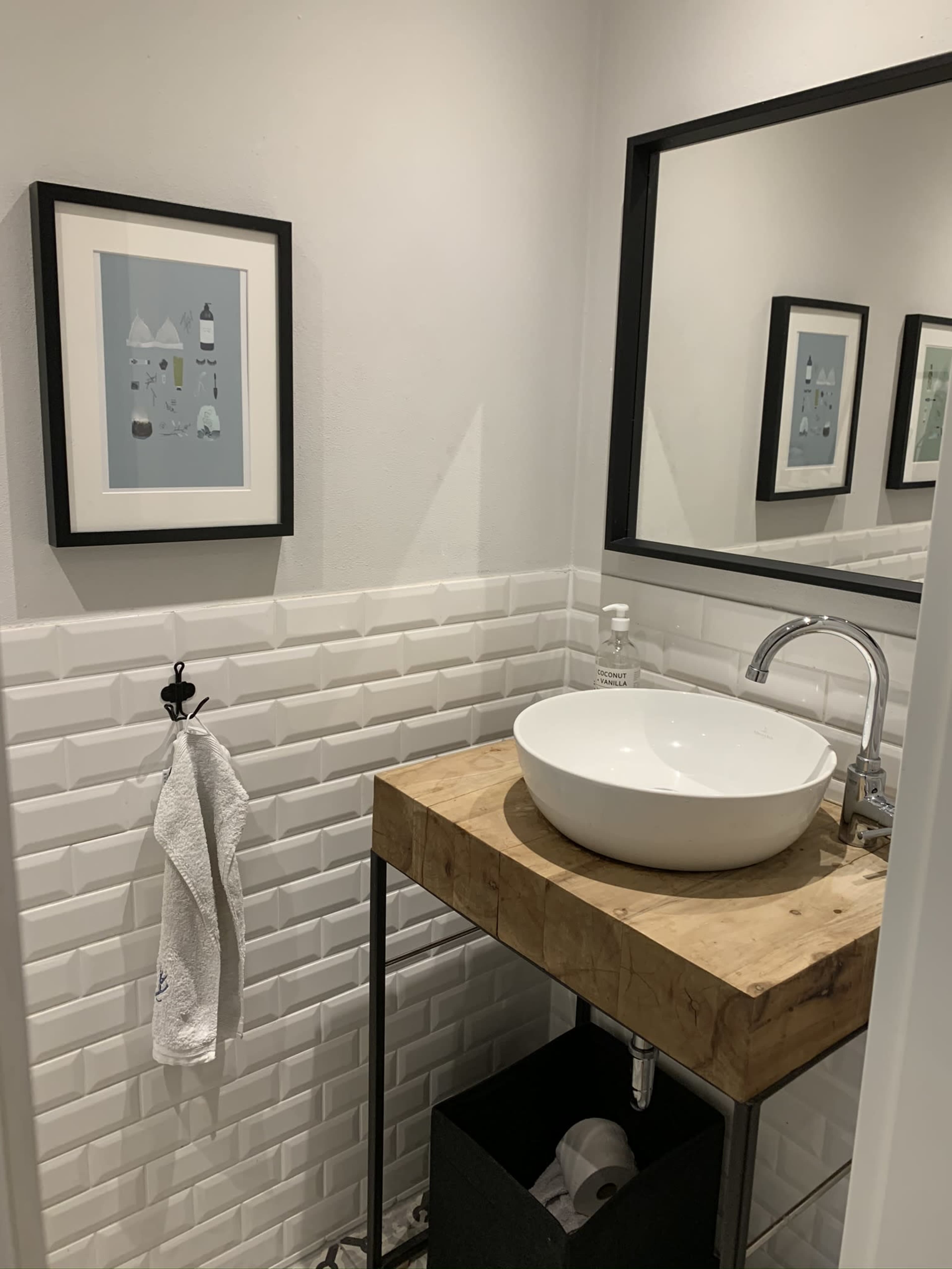 The image shows a modern bathroom with a white vessel sink on a wooden countertop, surrounded by tiled walls and two framed paintings.