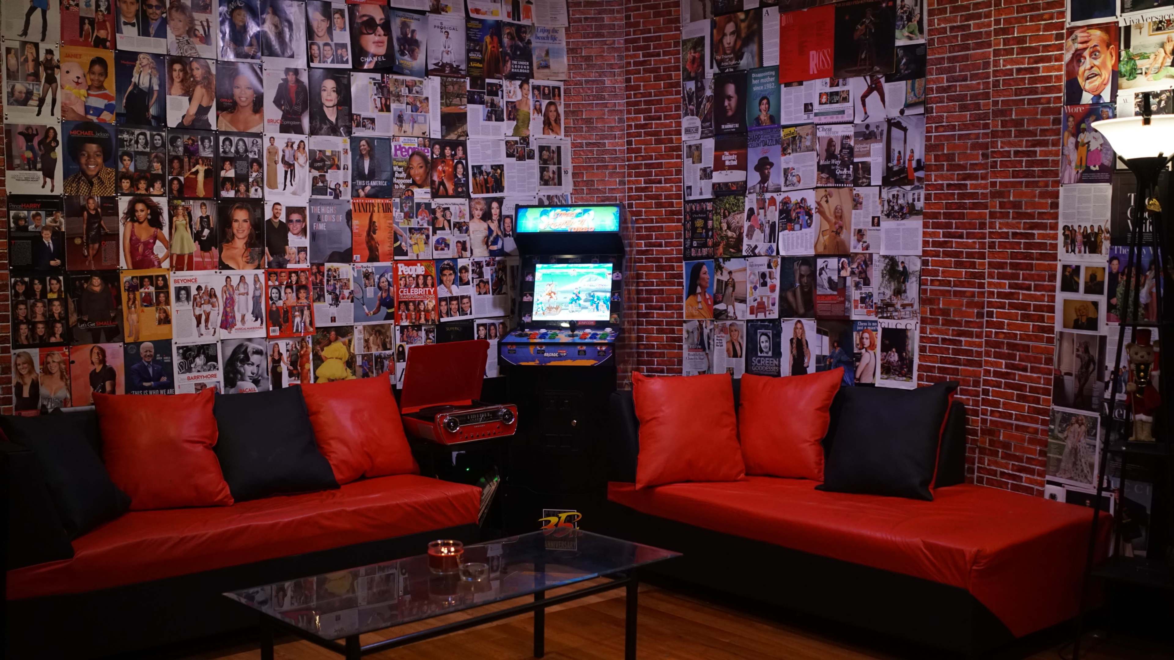 The room features two red couches facing a glass coffee table, surrounded by walls covered with various magazine clippings and photographs, with a vintage arcade machine in one corner.