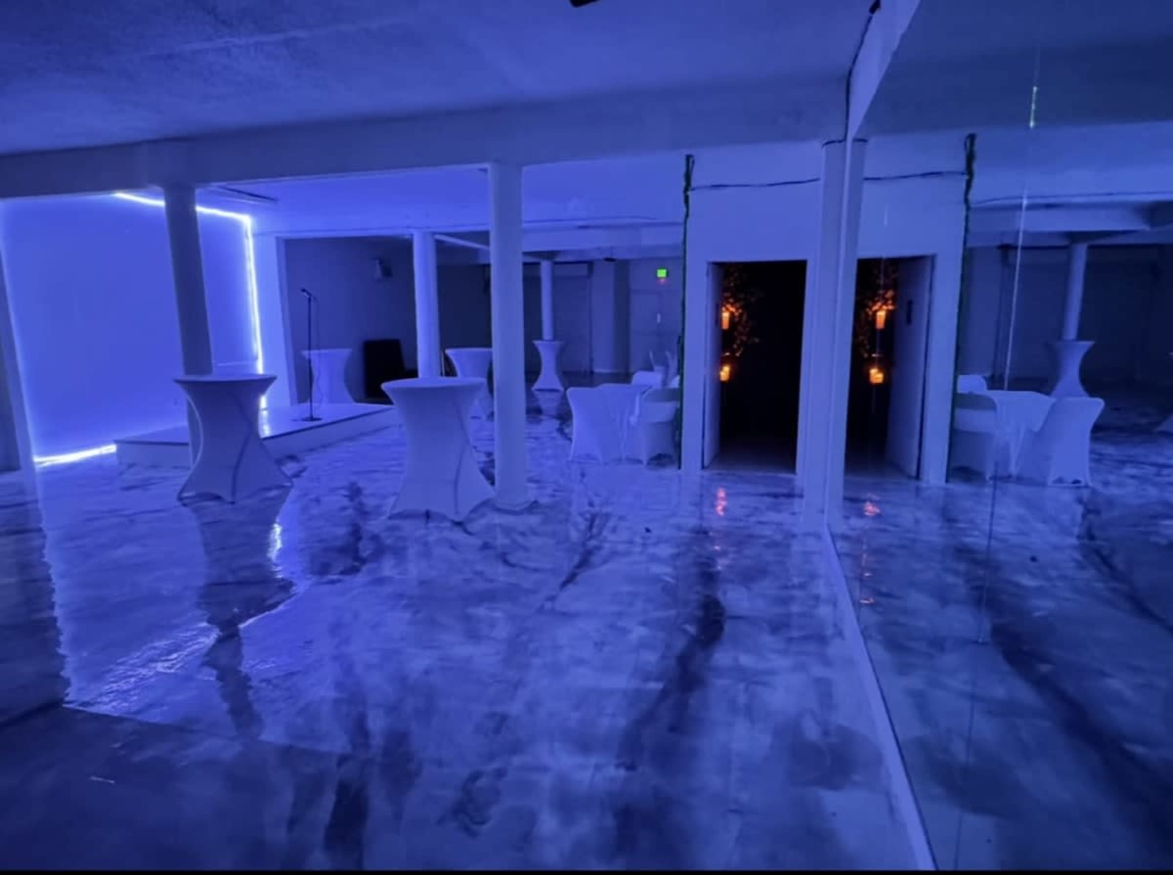 The image shows a dimly lit event space with white tables, mirrored walls, and blue lighting creating a modern atmosphere.